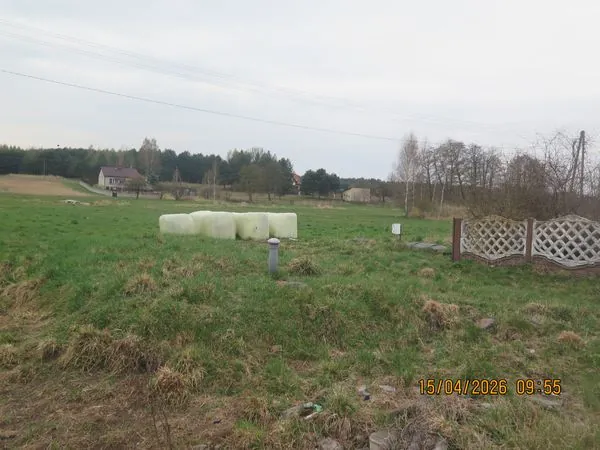 Krajobraz wiejski z zieloną łąką na pierwszym planie. W centrum znajdują się cztery okrągłe bele siana owinięte w białą folię. W tle widać grupę drzew, kilka domów mieszkalnych oraz linię wysokiego napięcia przecinającą niebo. W prawym dolnym rogu widnieje czerwony napis z datą i godziną: 15/04/2026 09:55.