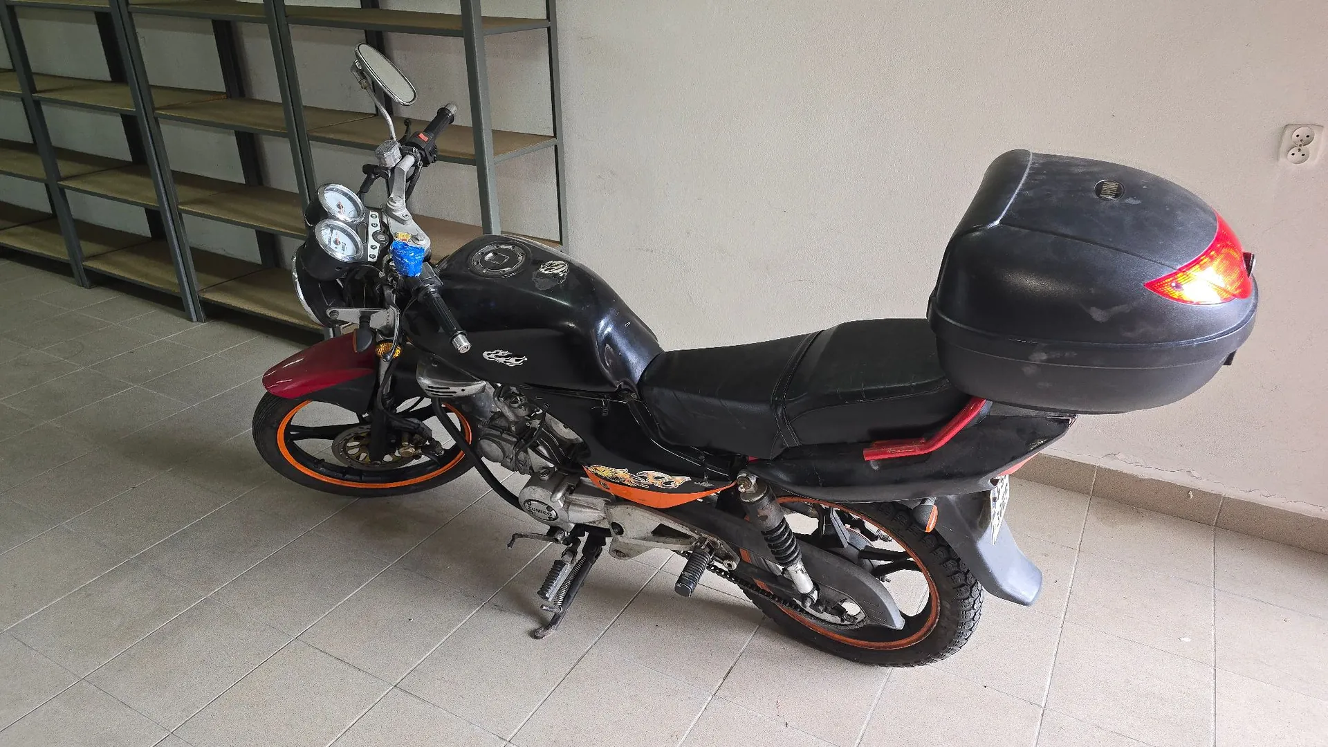 Czarny motocykl zaparkowany w pomieszczeniu na jasnej, kafelkowej podłodze. Pojazd posiada czarny bak, czarne siedzenie oraz zamontowany z tyłu czarny kufer. Felgi kół mają pomarańczowe obramowania. W tle widoczna jest biała ściana oraz metalowy regał z półkami.