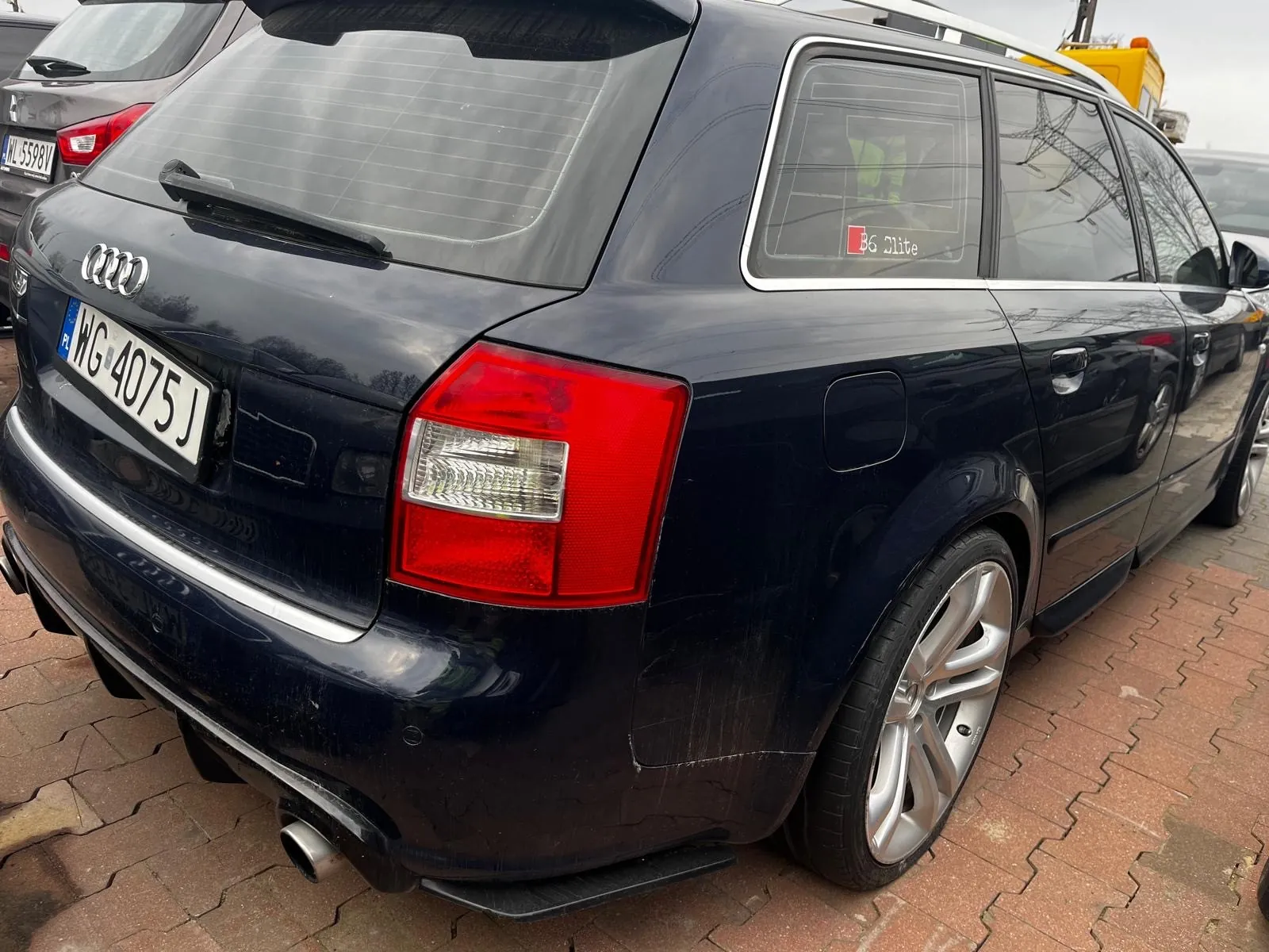 Czarny samochód Audi typu kombi z widoczną tablicą rejestracyjną WG 4075 J i naklejką B6 Elite na tylnej szybie. Pojazd zaparkowany na ceglanym placu, w tle inne samochody, w tym z tablicą NL 5598V. Niebo zachmurzone.