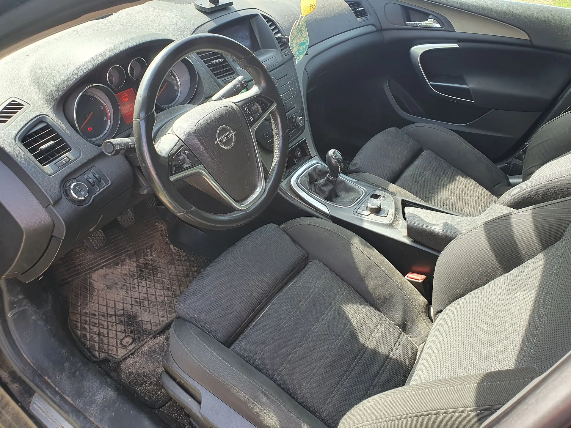 Wnętrze samochodu marki Opel widziane z perspektywy pasażera. Widoczna jest czarna kierownica z logo producenta, deska rozdzielcza z zegarami, konsola środkowa z drążkiem zmiany biegów oraz dwa przednie fotele obite ciemnoszarą tkaniną. Na podłodze znajduje się gumowa mata z wypustkami.