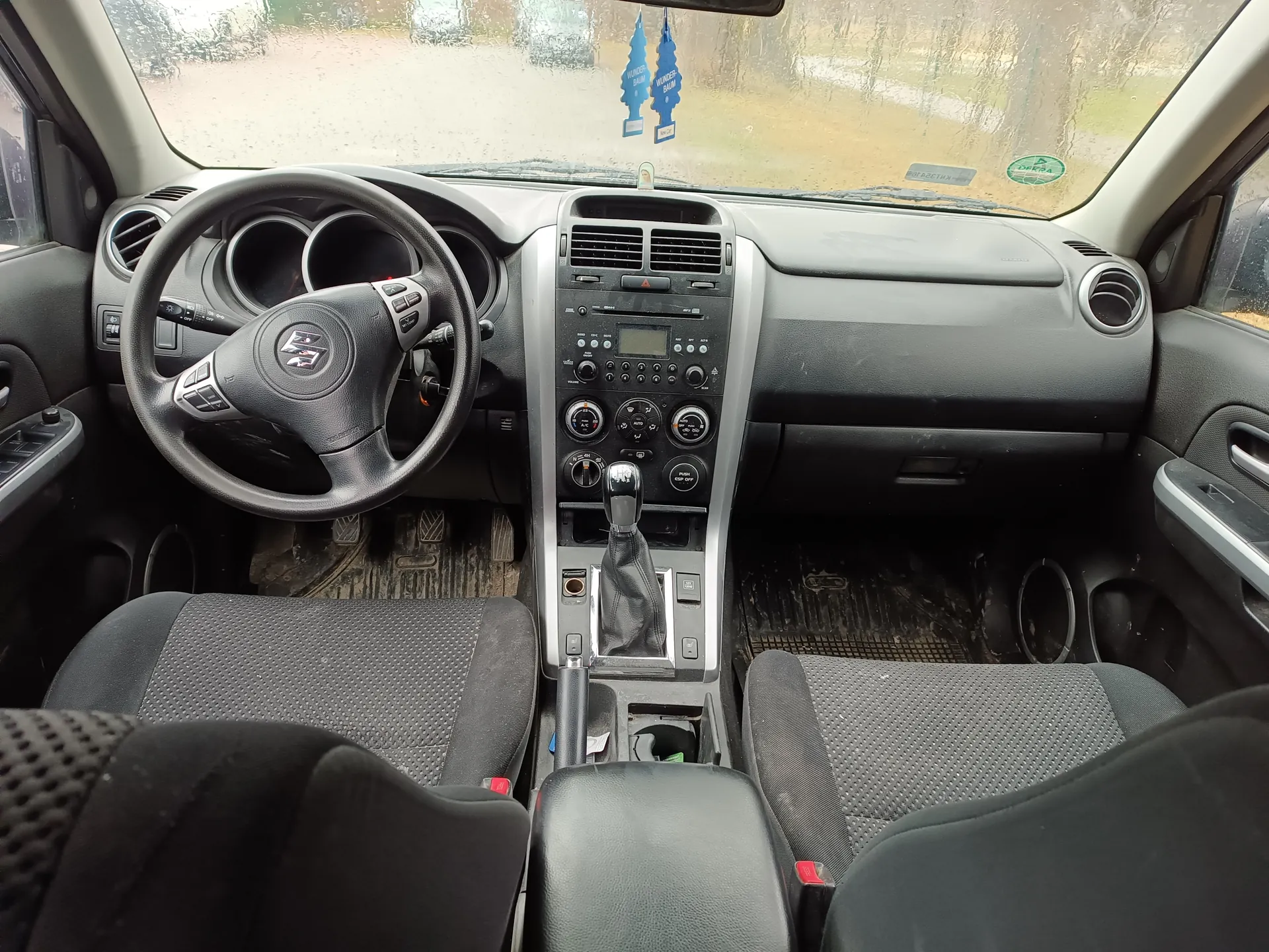 Wnętrze samochodu z widokiem na deskę rozdzielczą, kierownicę z logo, centralną konsolę z systemem audio i klimatyzacją, dźwignię zmiany biegów oraz siedzenia. Na przedniej szybie widoczne są krople deszczu, a na lusterku wstecznym wiszą dwa niebieskie odświeżacze powietrza w kształcie choinek. W tle przez szybę widać rozmyty krajobraz z drzewami i drogą. Deska rozdzielcza i elementy są czarno-szare, siedzenia mają siateczkowy wzór. Na przedniej szybie znajdują się również naklejki, w tym zielona okrągła.