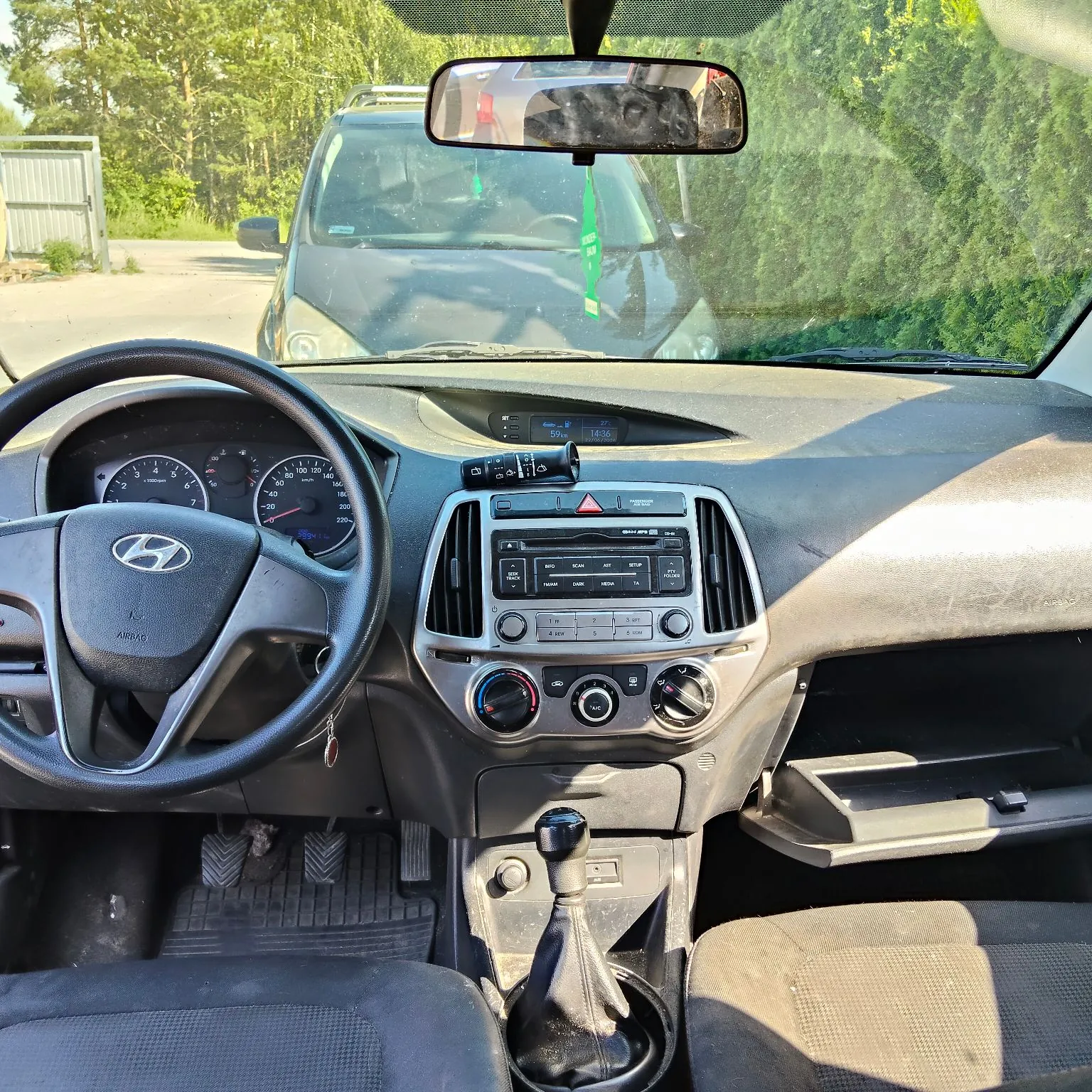 Wnętrze samochodu marki Hyundai, widok z perspektywy kierowcy. Widoczna jest czarna kierownica z logo Hyundai, deska rozdzielcza z radiem, klimatyzacją i licznikiem. W centralnej konsoli znajduje się lewarek zmiany biegów. Przez przednią szybę widać zaparkowany przed autem ciemny samochód oraz zielone drzewa w tle.