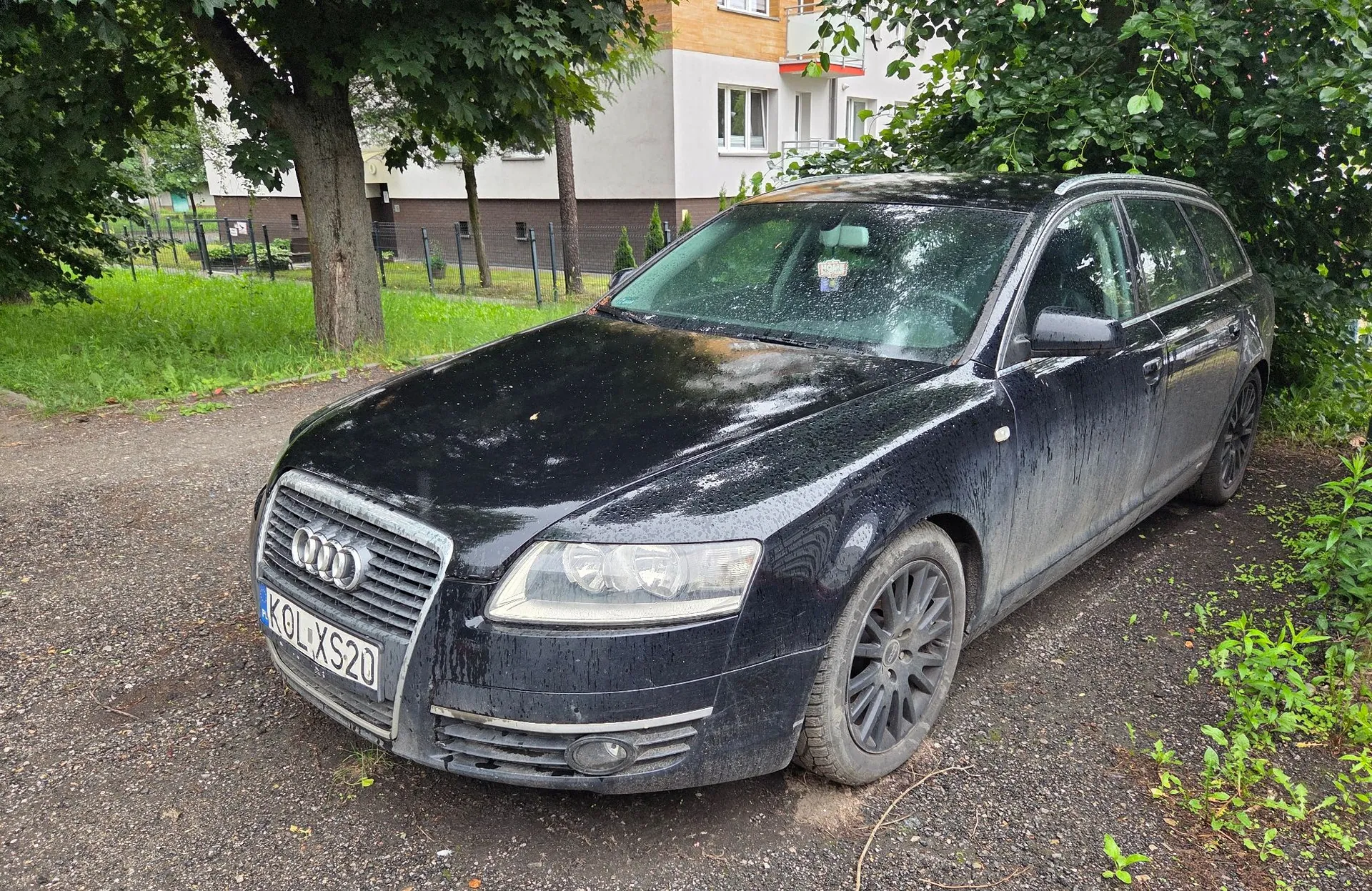 Czarny, zabrudzony samochód marki Audi w nadwoziu kombi, zaparkowany na żwirowym podłożzu obok trawnika i drzew. Na przodzie auta widoczna jest tablica rejestracyjna o numerze KOL XS20. W tle znajduje się biały budynek mieszkalny z oknami.