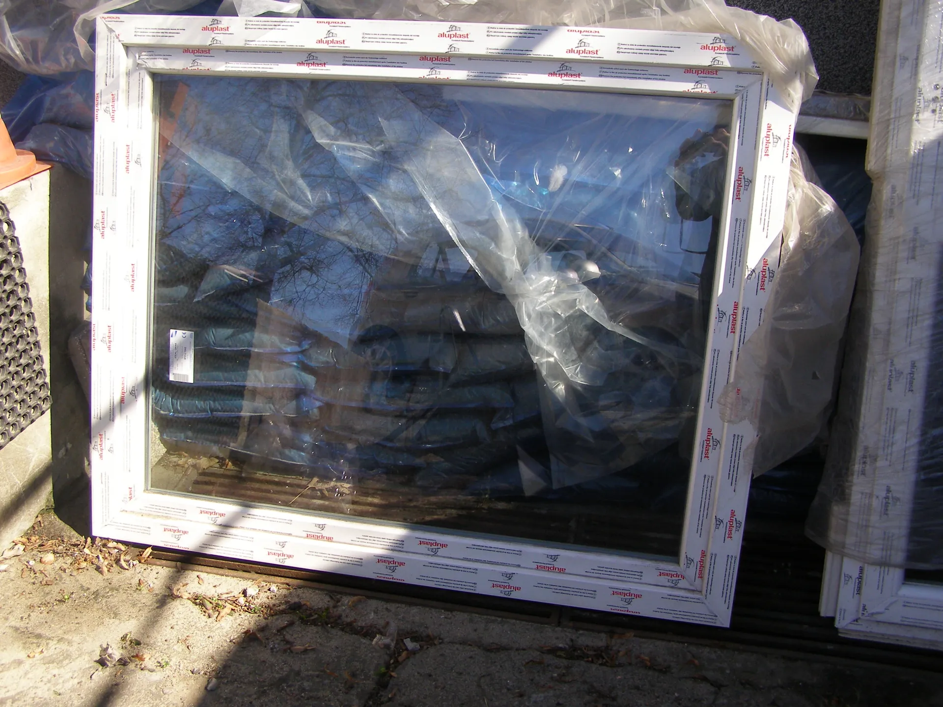 Biały aluminiowy rama okienna z nadrukiem 'aluplast' na białym tle z czerwonymi literami, owinięta przezroczystą folią. W tle widoczne są stosy materiałów, liście na ziemi i fragmenty innych opakowań. Na szybie znajduje się biała etykieta z nieczytelnym tekstem.