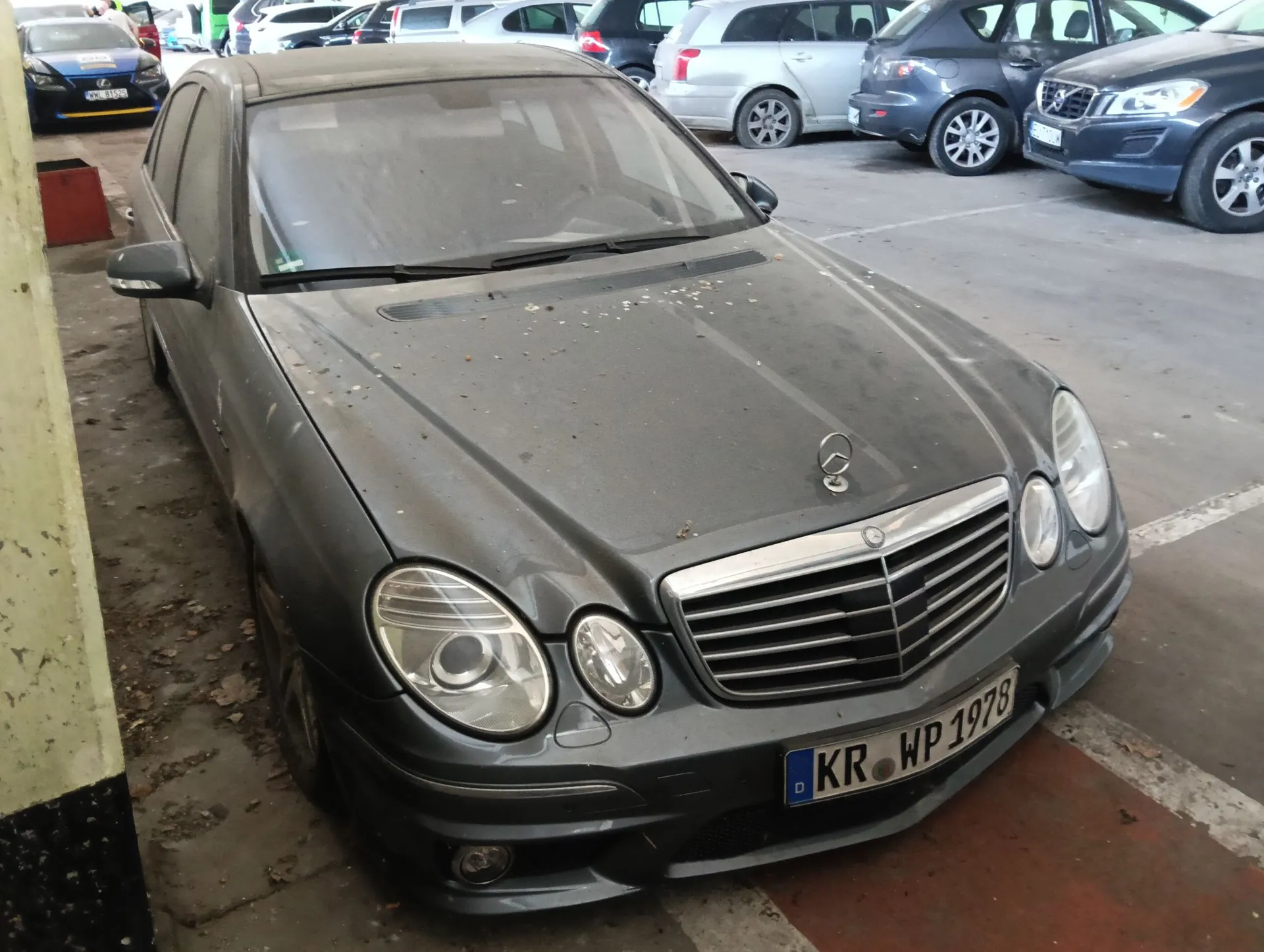 Szary samochód osobowy marki Mercedes-Benz zaparkowany na parkingu. Pojazd jest zakurzony i ma na masce kilka suchych liści. Widoczna jest tablica rejestracyjna o treści KR WP 1978. W tle znajdują się inne zaparkowane samochody w różnych kolorach.