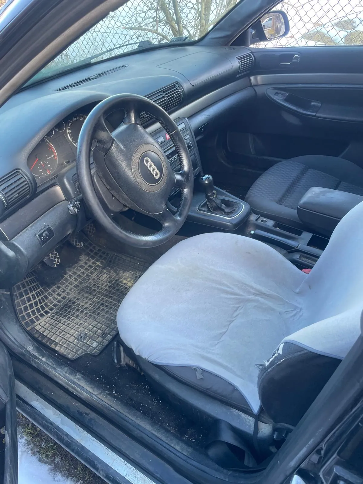 Wnętrze samochodu Audi z kierownicą z logo, manualną skrzynią biegów, czarnymi siedzeniami i szarą pokrywą na fotelu kierowcy. Brudne dywany, otwarte drzwi, widok na siatkę i śnieg na zewnątrz. Tablica rozdzielcza z licznikami, przyciski klimatyzacji.
