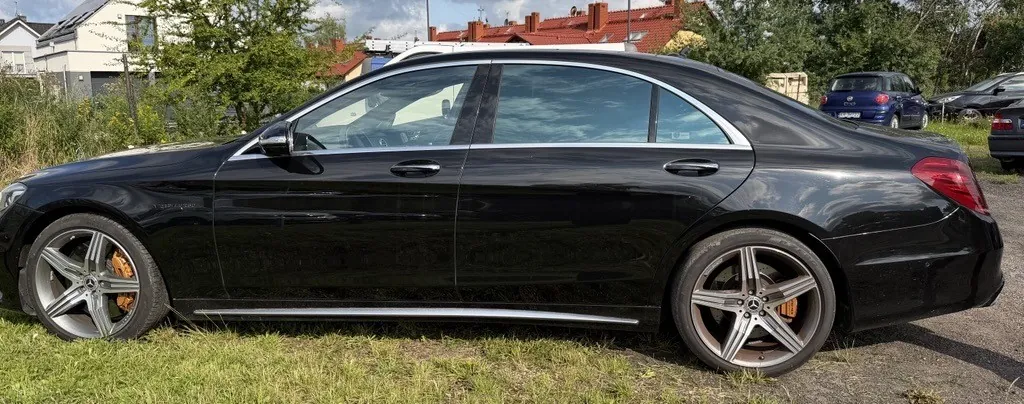 Czarny samochód osobowy marki Mercedes-Benz zaparkowany bokiem na trawie i asfalcie. Pojazd posiada srebrne felgi z pięcioma ramionami i widoczne pomarańczowe zaciski hamulcowe. W tle widać zielone drzewa, krzewy oraz dachy domów mieszkalnych.