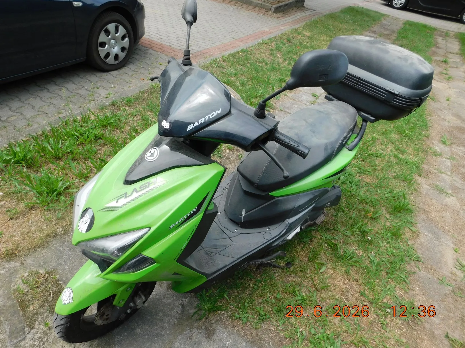Jasnozielony skuter marki Barton model Flash z czarnym kufrem z tyłu, zaparkowany na trawie i kostce brukowej. Na przedniej owiewce widnieje napis BARTON, a na boku napis FLASH i BARTON. W prawym dolnym rogu zdjęcia znajduje się pomarańczowy znacznik daty i godziny: 29.6.2026 12:36.