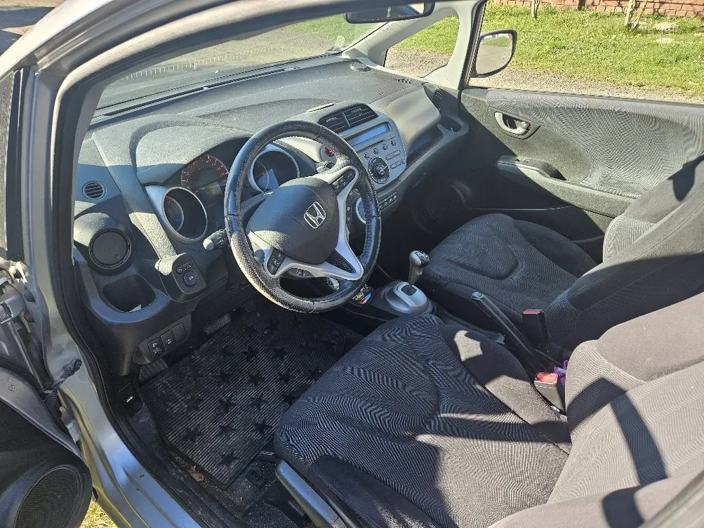 Wnętrze samochodu marki Honda z otwartą drzwiami od strony kierowcy. Widoczna kierownica z logo Hondy, deska rozdzielcza z licznikami, czarne siedzenia tapicerowane tkaniną, dźwignia zmiany biegów oraz czarny dywanik podłogowy z wzorem gwiazdek. Poza samochodem widoczna trawa i ceglana ściana.