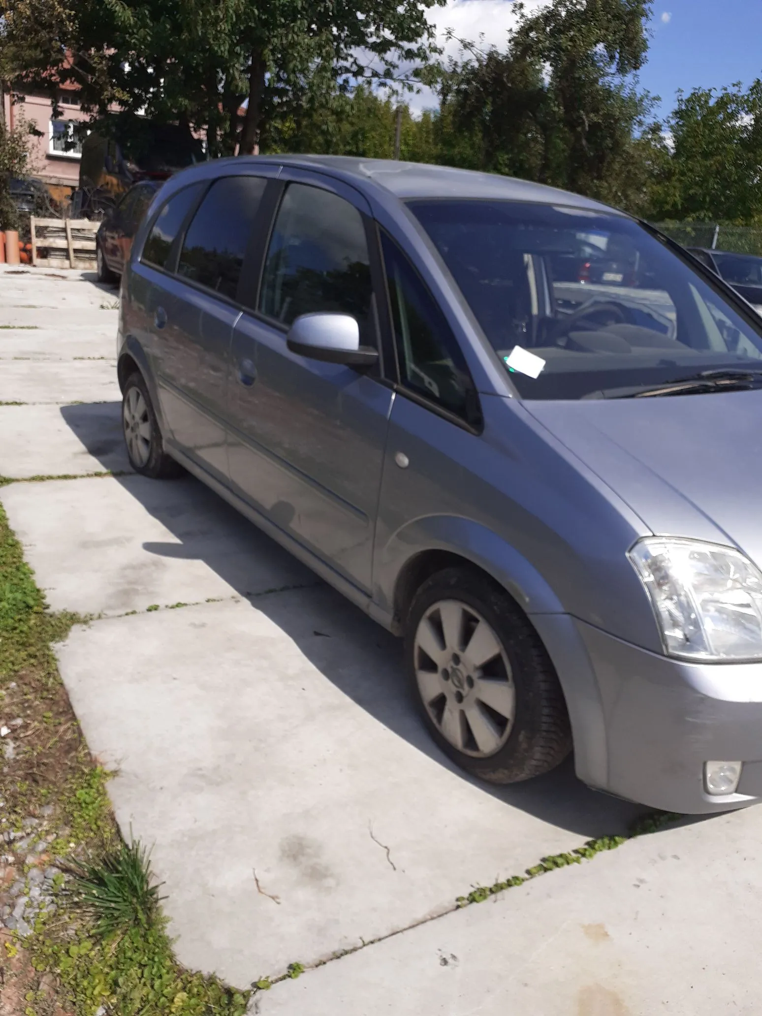 Srebrny samochód typu minivan zaparkowany na betonowym podjeździe. Pojazd jest pokazany z perspektywy przedniego lewego boku. W tle widoczne są zielone drzewa oraz fragment budynku.