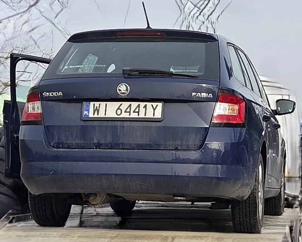 Tył niebieskiego samochodu marki Škoda Fabia z otwartą lewą tylną drzwiami. Widoczna tablica rejestracyjna z polskim kodem PL i numerem WI 6441Y. Samochód jest zabrudzony, szczególnie dolna część. W tle suche drzewa i fragment innego pojazdu. Na bagażniku widoczne logo marki oraz napisy ŠKODA i FABIA.