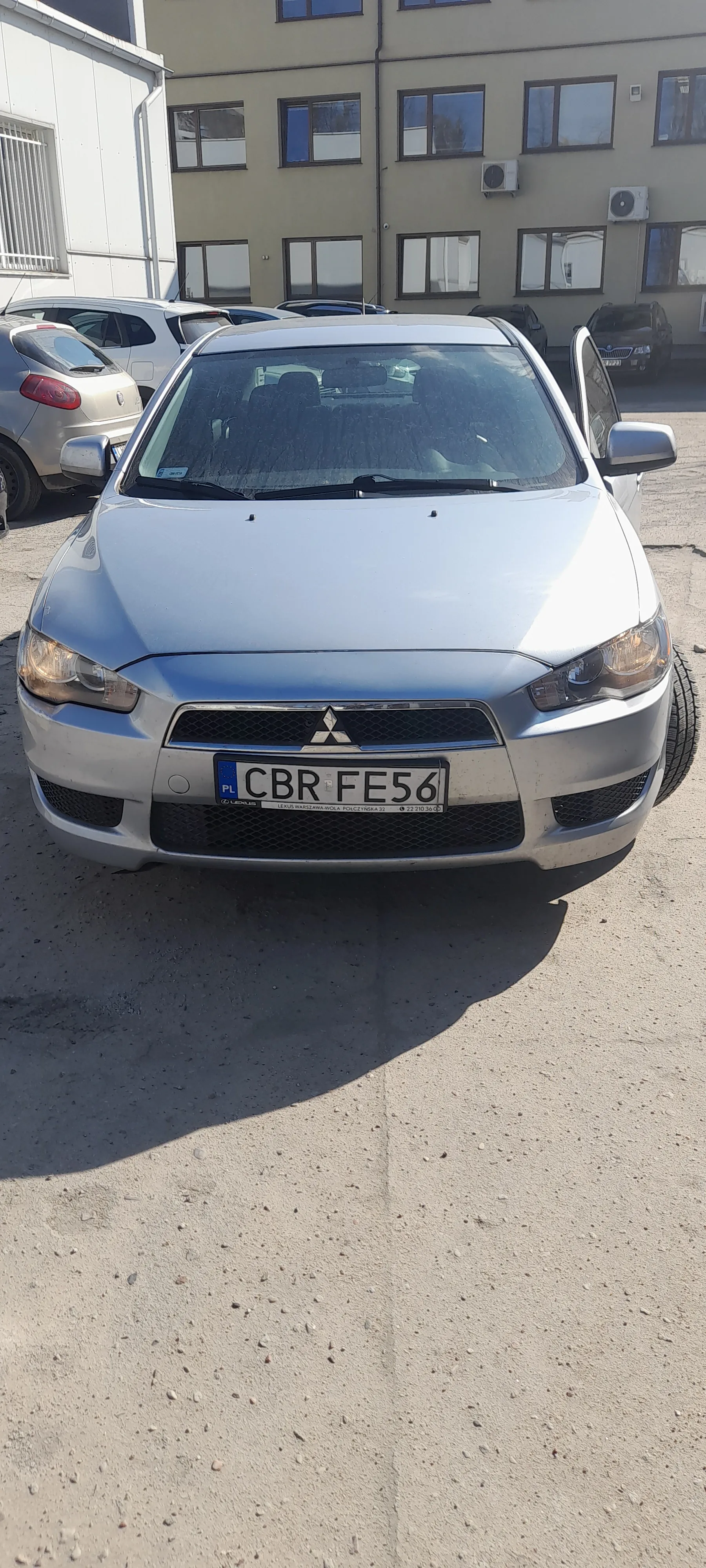 Srebrny samochód marki Mitsubishi z tablicą rejestracyjną CBR FE56 zaparkowany na asfaltowej powierzchni. W tle widoczny budynek z oknami i klimatyzatorami oraz częściowo widoczny inny samochód.
