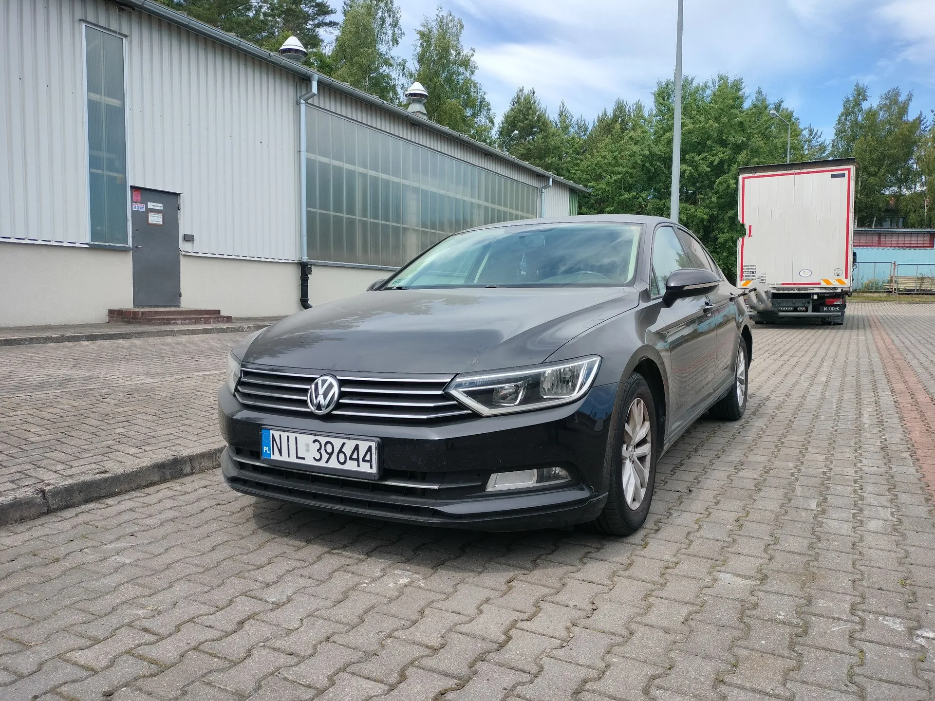 Czarny samochód marki Volkswagen zaparkowany na kostce brukowej przed jasnym, przemysłowym budynkiem z dużymi oknami. Samochód ma tablicę rejestracyjną o numerze NIL 39644. W tle po prawej stronie widoczna jest biała naczepa ciężarówki oraz zielone drzewa pod błękitnym niebem z chmurami.