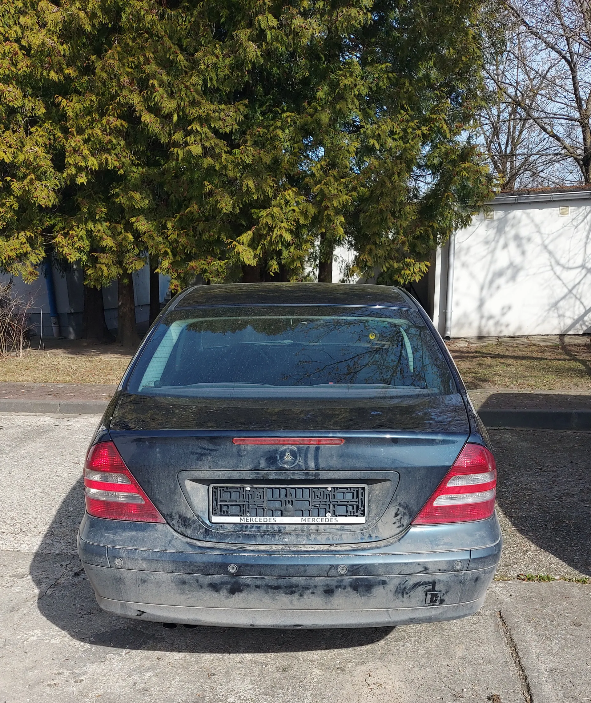 Tył brudnego samochodu marki Mercedes klasy E, zaparkowanego na betonowej powierzchni. Widoczne są czerwone światła tylnego, zanieczyszczony bagażnik oraz tablica rejestracyjna z ramką z napisem "MERCEDES" na górze i dole. W tle drzewa z zielonymi liśćmi, biały budynek oraz jasne niebo. Samochód ma ciemnoniebieską lub szarą barwę, a na powierzchni widoczne są plamy kurzu i brudu.
