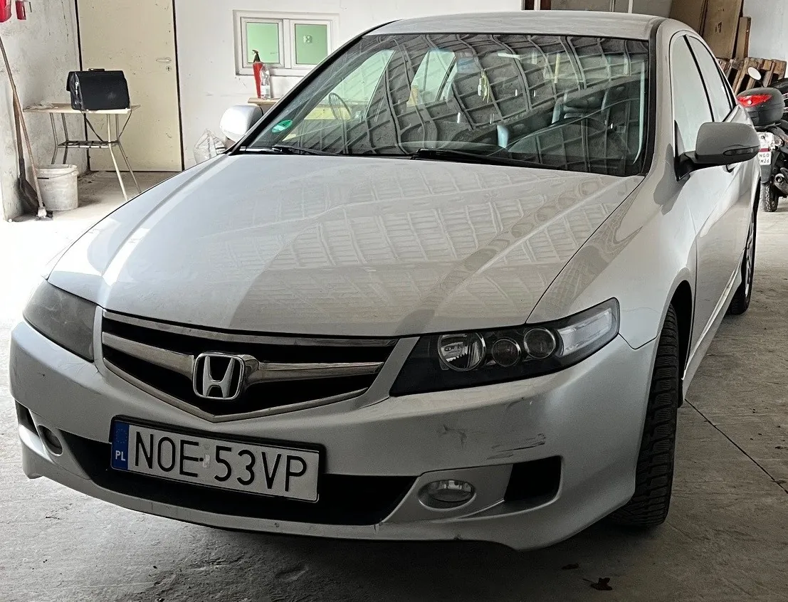 Srebrny samochód marki Honda z tablicą rejestracyjną PL NOE 53VP, zaparkowany w garażu. W tle widoczne są stół z czarną torbą, gaśnica, okna z zielonymi szybami oraz motocykl. Podłoga garażu jest betonowa, a na ścianie znajdują się narzędzia i pojemniki.