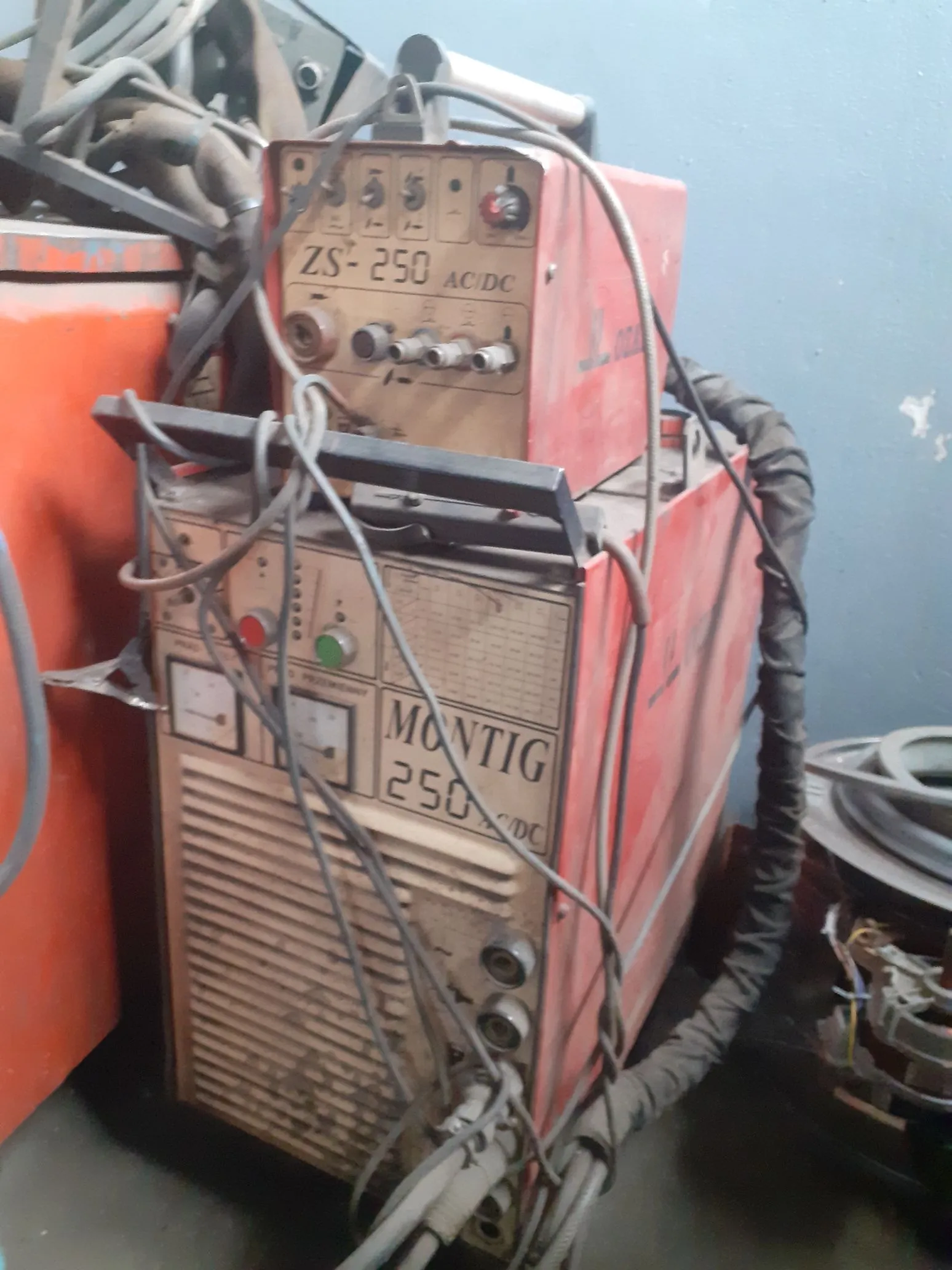 Dwa czerwone, przemysłowe urządzenia spawalnicze ustawione jedno na drugim. Górne urządzenie posiada napis 'ZS-250 AC/DC' oraz panel z pokrętłami i przełącznikami. Dolne urządzenie ma napis 'MONTIG 250 DC', panel sterowania z zielonym i czerwonym przyciskiem oraz dużą kratownicę wentylacyjną z przodu. Oba urządzenia są oplecione szarymi kablami i wężami.