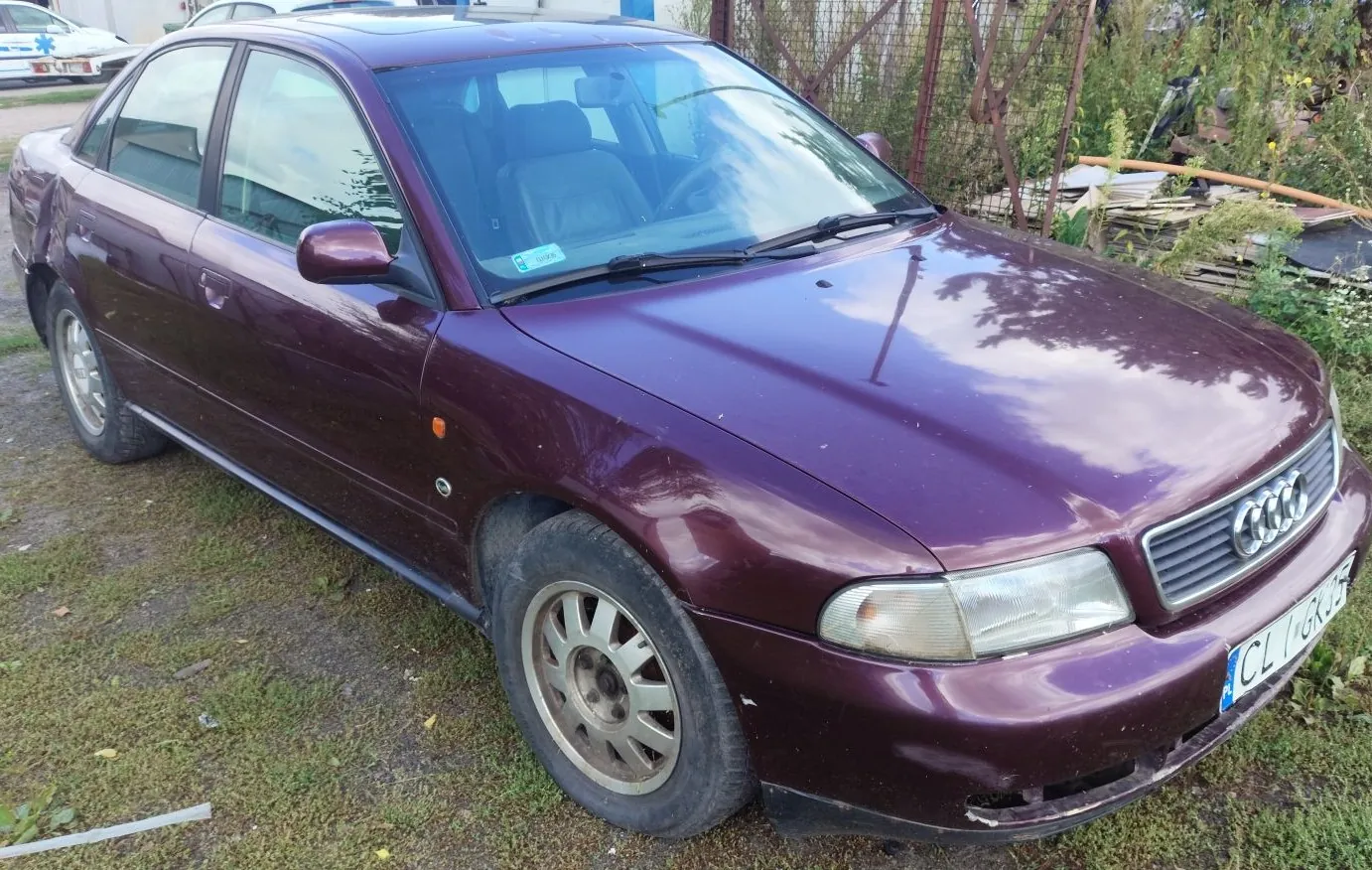 Bordowy samochód marki Audi w nadwoziu sedan, zaparkowany na trawiastym podłożu. Widoczny jest przód i lewy bok pojazdu. Samochód posiada tablicę rejestracyjną o numerze CL 6GK5. W tle znajduje się metalowy płot oraz roślinność.