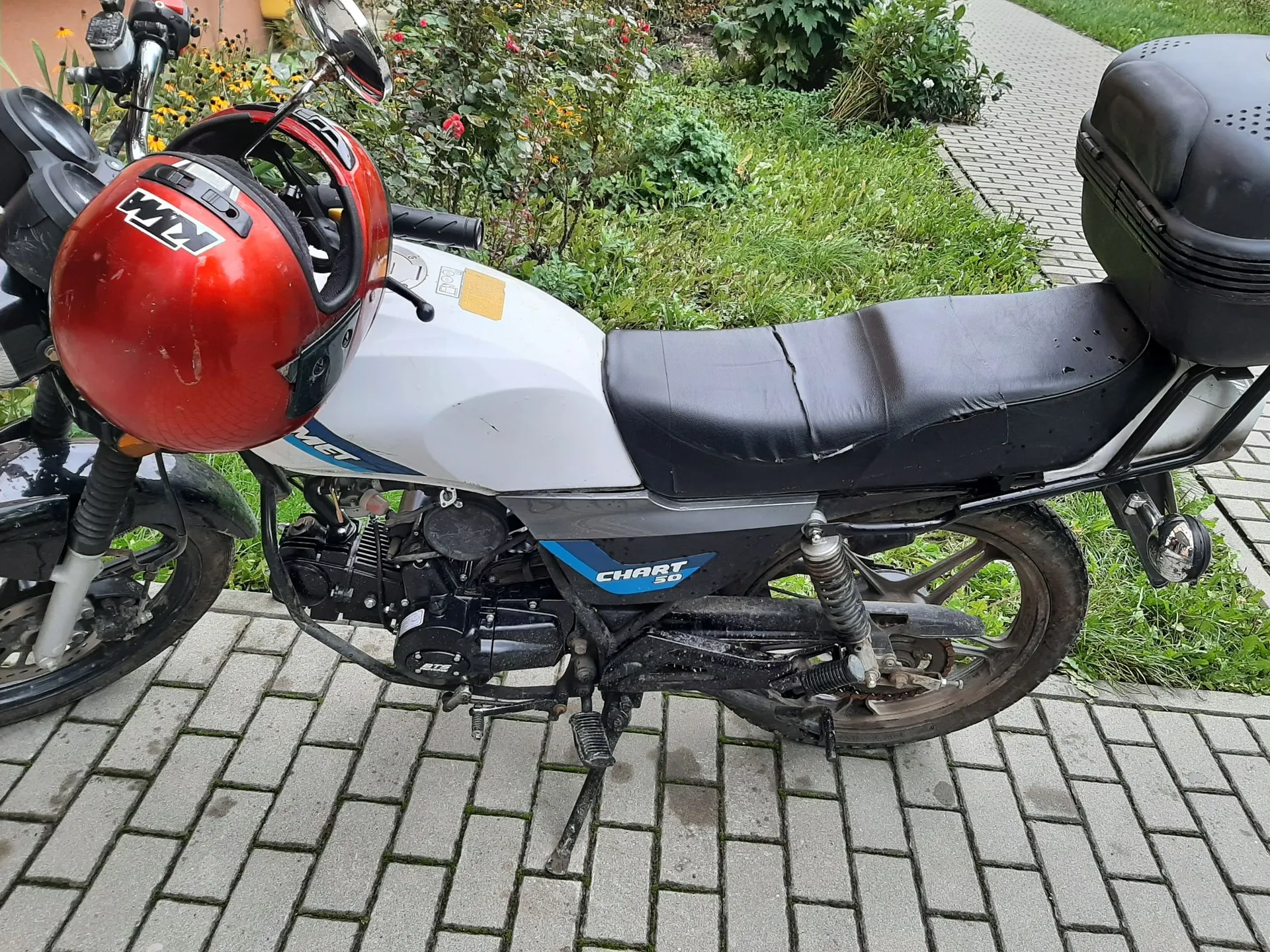 Biały motocykl marki Chart 50 zaparkowany na kostce brukowej. Na kierownicy znajduje się czerwony kask z białym napisem 'KW'. Motocykl posiada czarne siedzenie oraz czarny kufer z tyłu. W tle widoczna jest trawa i krzewy.