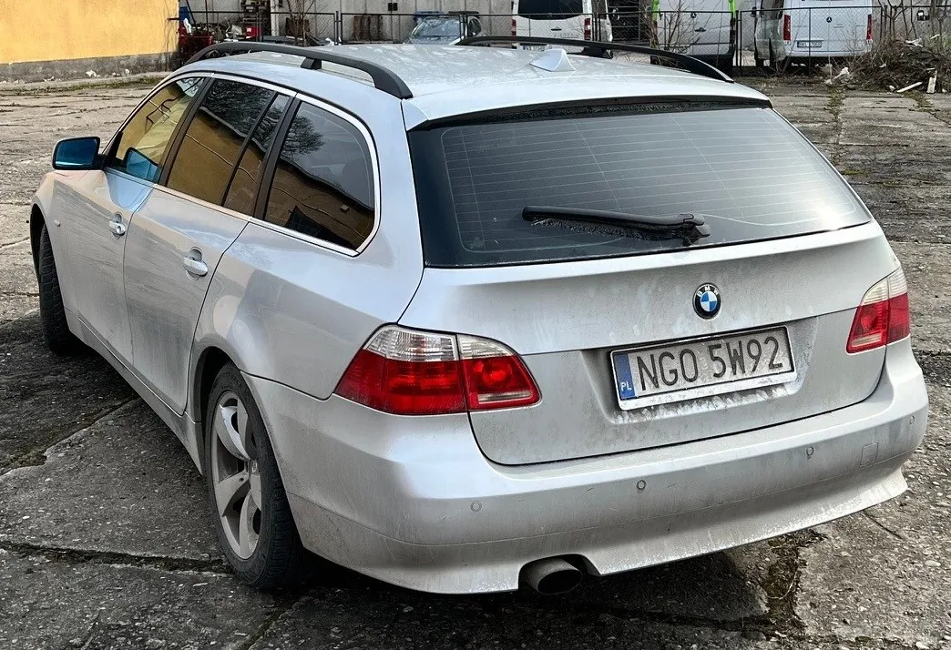 Srebrny samochód osobowy typu kombi marki BMW, widok od tyłu. Na tylnej szybie wycieraczka, a na bagażniku logo BMW. Tablica rejestracyjna z napisem PL NGO 5W92. Samochód stoi na betonowej powierzchni z widocznymi pęknięciami. W tle żółta ściana budynku, ogrodzenie, inne pojazdy i suche krzewy.