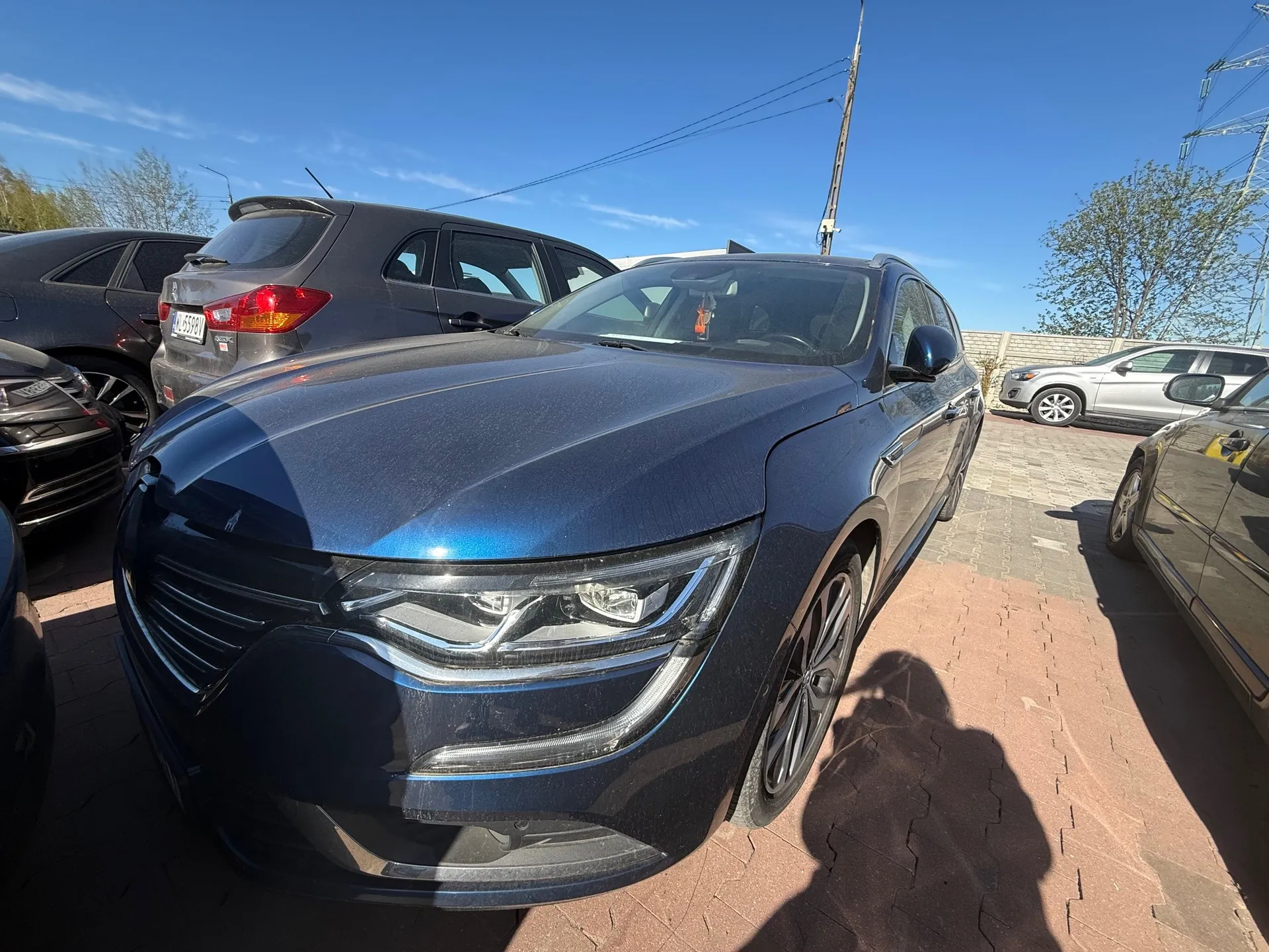 Ciemnoniebieski samochód osobowy na pierwszym planie, zaparkowany na ceglanym placu. W tle widoczne są inne pojazdy: szary SUV z tablicą rejestracyjną WU 5528V, czarny samochód po lewej stronie oraz srebrny samochód w oddali. Tło stanowi jasnoniebieskie niebo, drzewa i linie energetyczne. Cień osoby fotografującej widoczny na nawierzchni.