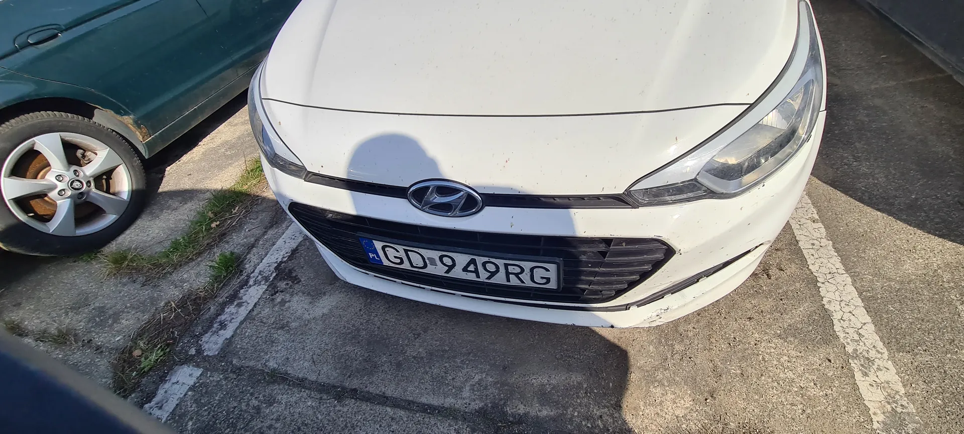 Przednia część białego samochodu marki Hyundai z tablicą rejestracyjną PL GD 949 RG. Po lewej stronie widoczna część zielonego samochodu z kołem. Samochody zaparkowane na asfaltowym parkingu z białymi liniami i fragmentami trawy w szczelinach.