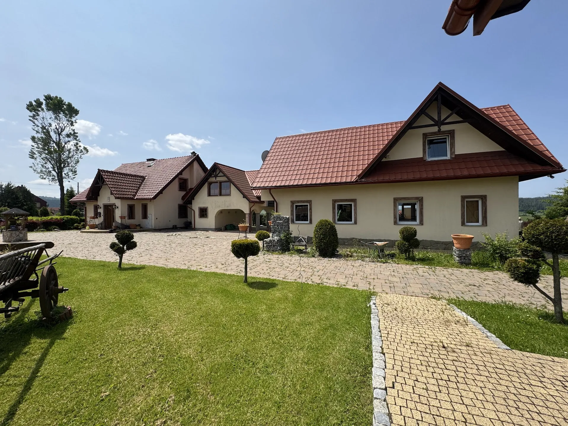 Widok na duży dom z czerwoną dachówką i beżowymi ścianami. Dom posiada kilka okien oraz garaż. Przed domem znajduje się brukowana podjazdowa aleja, zielony trawnik, przycięte krzewy oraz stary drewniany wóz na lewej stronie. W tle widać drzewa, błękitne niebo z kilkoma chmurami oraz fragmenty innych budynków. Na prawo od domu umieszczono donicę z rośliną na kamiennym postumencie.