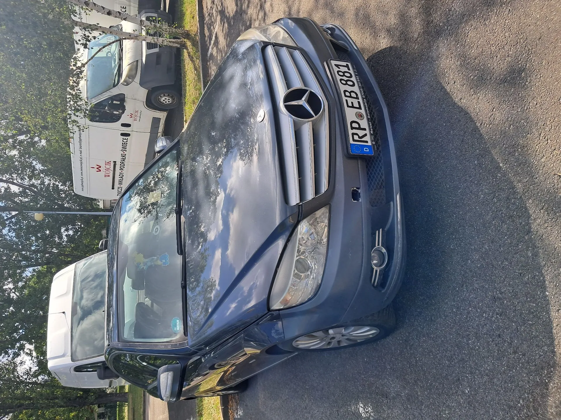Srebrny samochód marki Mercedes-Benz zaparkowany na asfaltowej nawierzchni. Widoczny jest przód pojazdu z charakterystycznym logo na grillu oraz tablicą rejestracyjną o numerze RP EB 881. W tle znajduje się biały samochód dostawczy z logo firmy 'WOCICKI' oraz zielone drzewa.