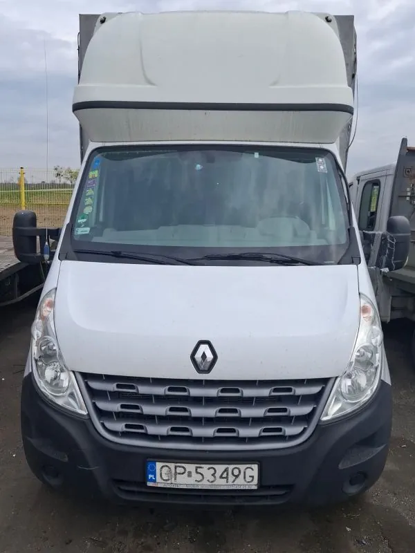 Biały samochód dostawczy marki Renault z widocznym logo na masce. Na przednim zderzaku umieszczona tablica rejestracyjna z napisem OP-5349G oraz oznaczeniem PL po lewej stronie. Przednią szybę zdobią naklejki po lewej stronie. W tle widoczne są inne pojazdy, żółte ogrodzenie i chmurne niebo. Samochód stoi na nierównym, brudnym podłożu.