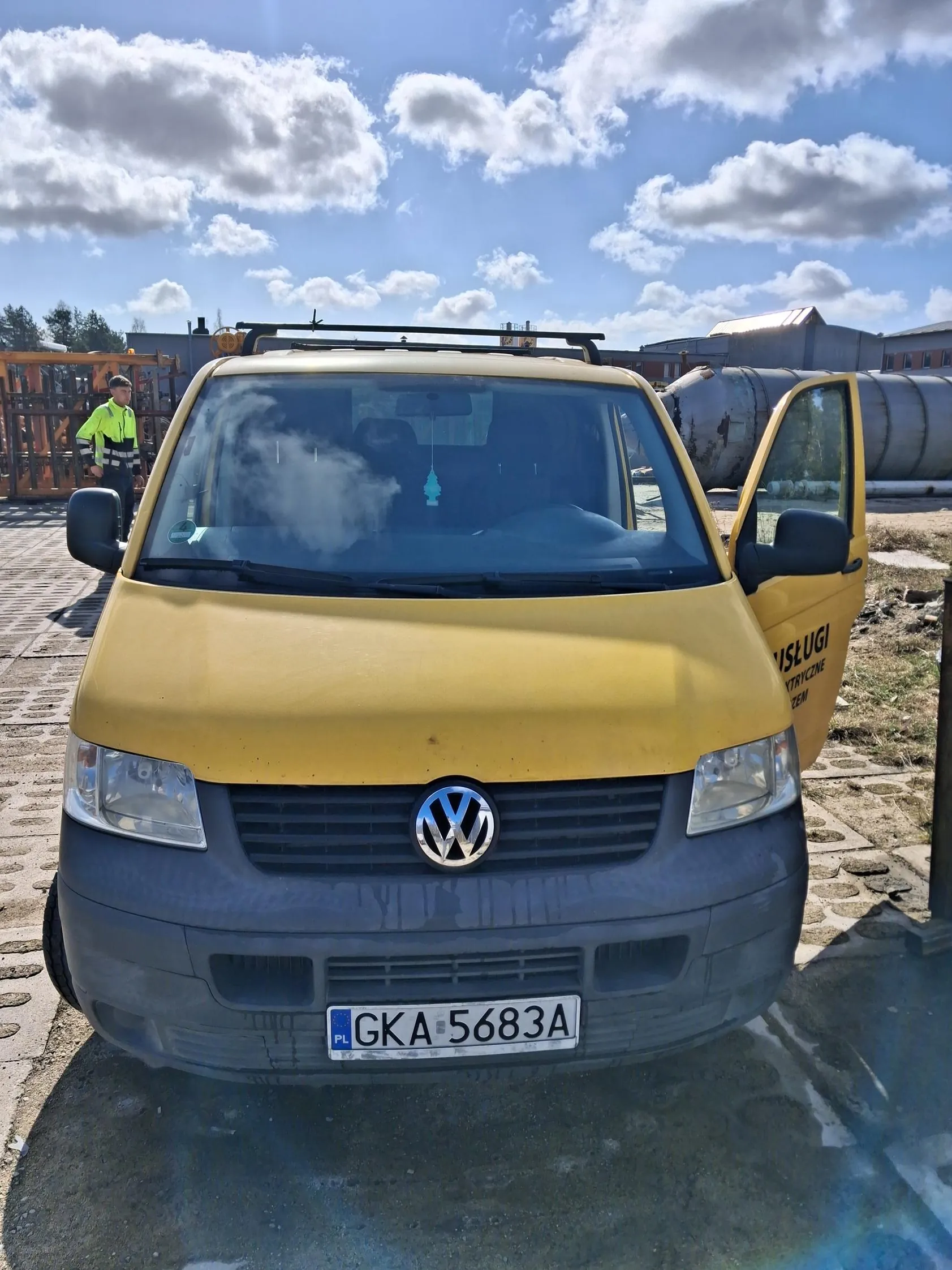 Żółto-szary samochód dostawczy marki Volkswagen z otwartą prawą drzwiami, tablicą rejestracyjną PL GKA 5683A i logo VW na masce. W tle osoba w jasnozielonej kurtce odblaskowej, przemysłowe zbiorniki, budynki i niebo z białymi chmurami. Samochód stoi na brukowanej powierzchni.