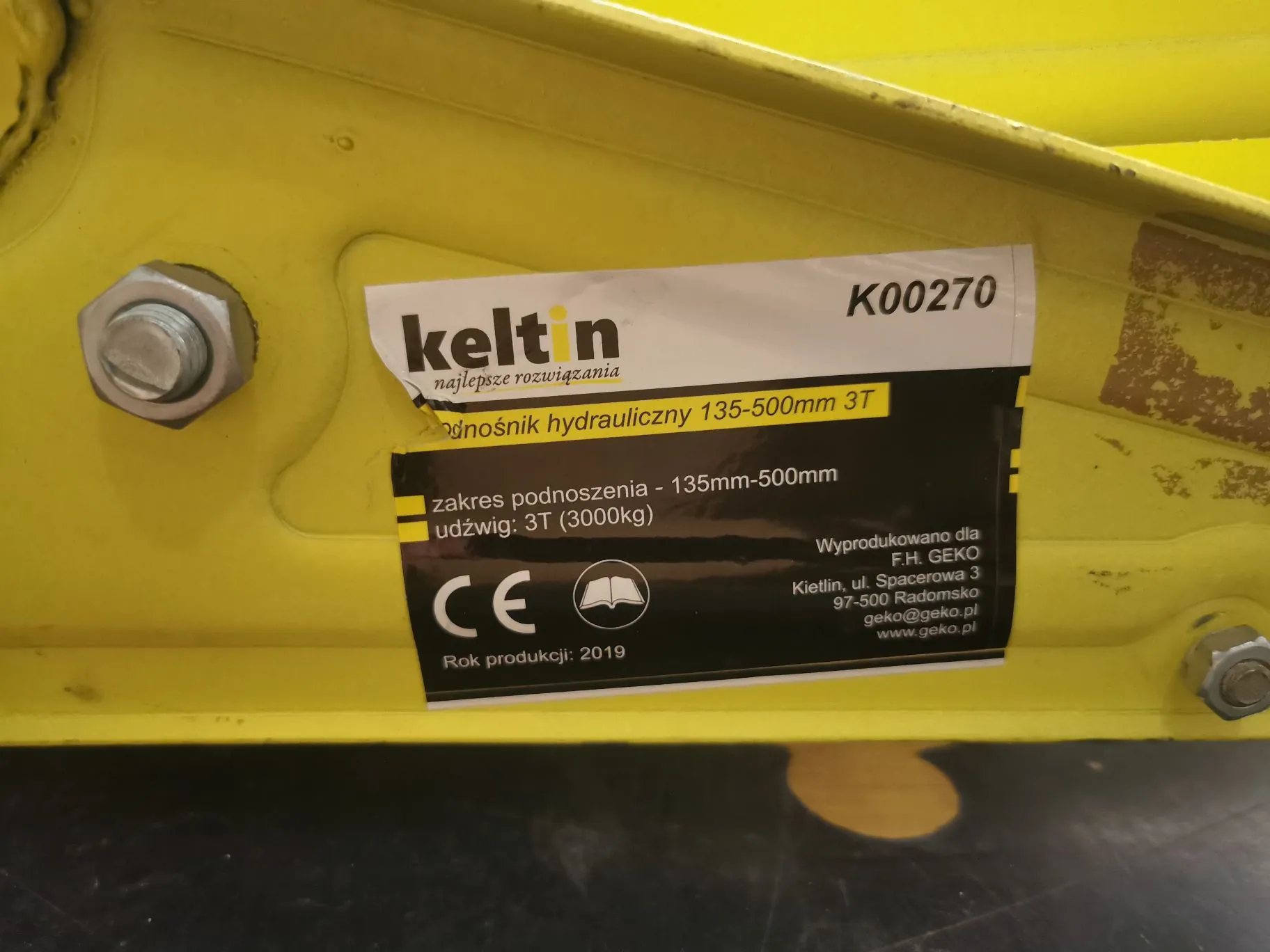 Zbliżenie na żółtą, metalową konstrukcję z przyklejoną biało-czarną etykietą informacyjną. Na etykiecie znajduje się logo 'keltin' z dopiskiem 'najlepsze rozwiązania' oraz numer 'K00270'. Tekst na etykiecie głosi: 'podnośnik hydrauliczny 135-500mm 3T', 'zakres podnoszenia - 135mm-500mm', 'udźwig: 3T (3000kg)'. Widoczne jest oznaczenie CE, ikona otwartej książki oraz informacja o producencie: 'Wyprodukowano dla: F.H. GEKO, Kietlin, ul. Spacerowa 3, 97-500 Radomsko, geko@geko.pl, www.geko.pl'. Na dole widnieje napis 'Rok produkcji: 2019'. Po lewej stronie etykiety znajduje się metalowa śruba z nakrętką.