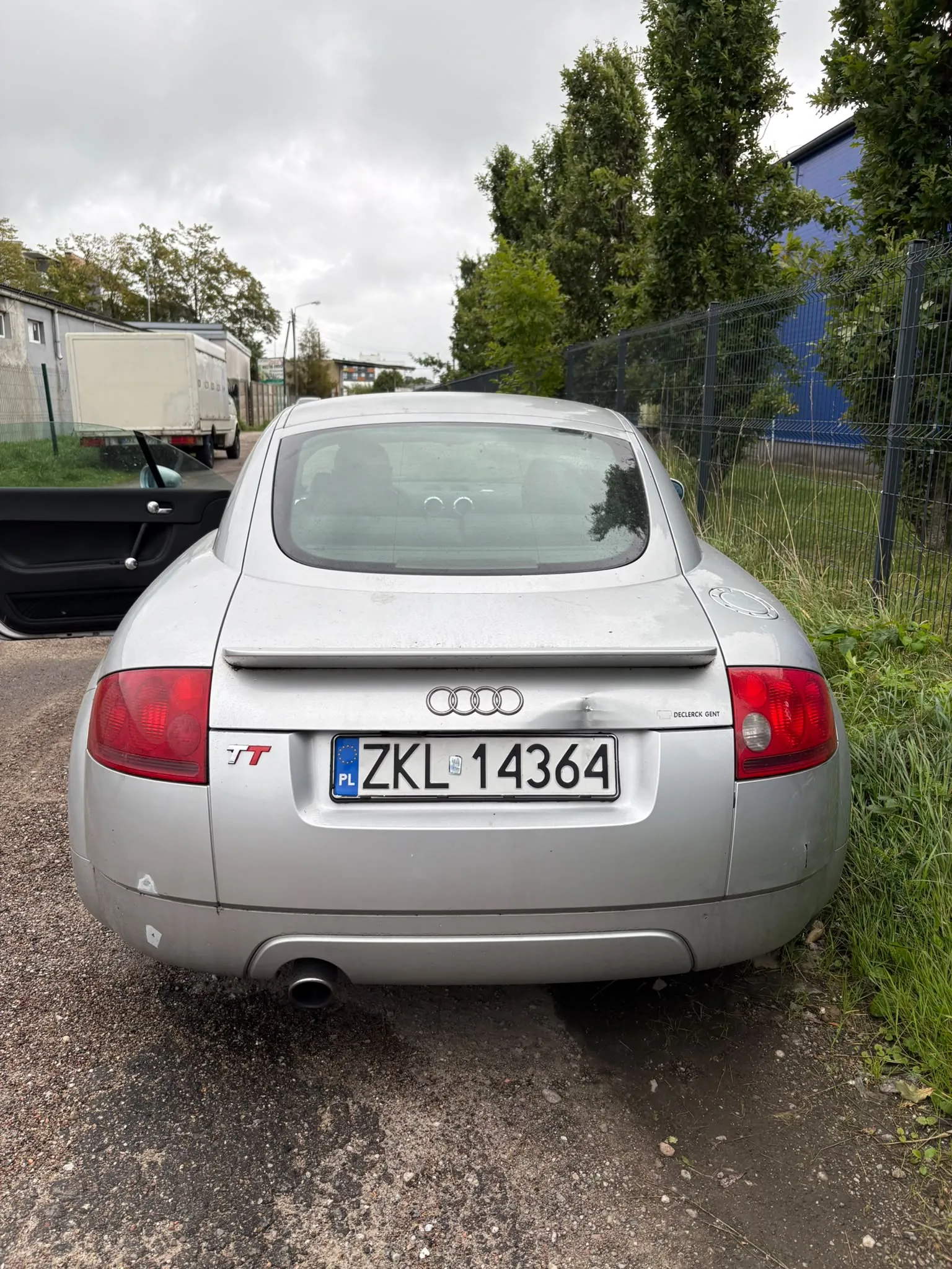Srebrny samochód Audi TT widziany od tyłu, zaparkowany na żwirowej drodze obok zielonego ogrodzenia i drzew. Na klapie bagażnika znajduje się logo Audi oraz polska tablica rejestracyjna o numerze ZKL 14364. Po lewej stronie widoczny jest fragment czarnego elementu nadwozia leżącego na ziemi.