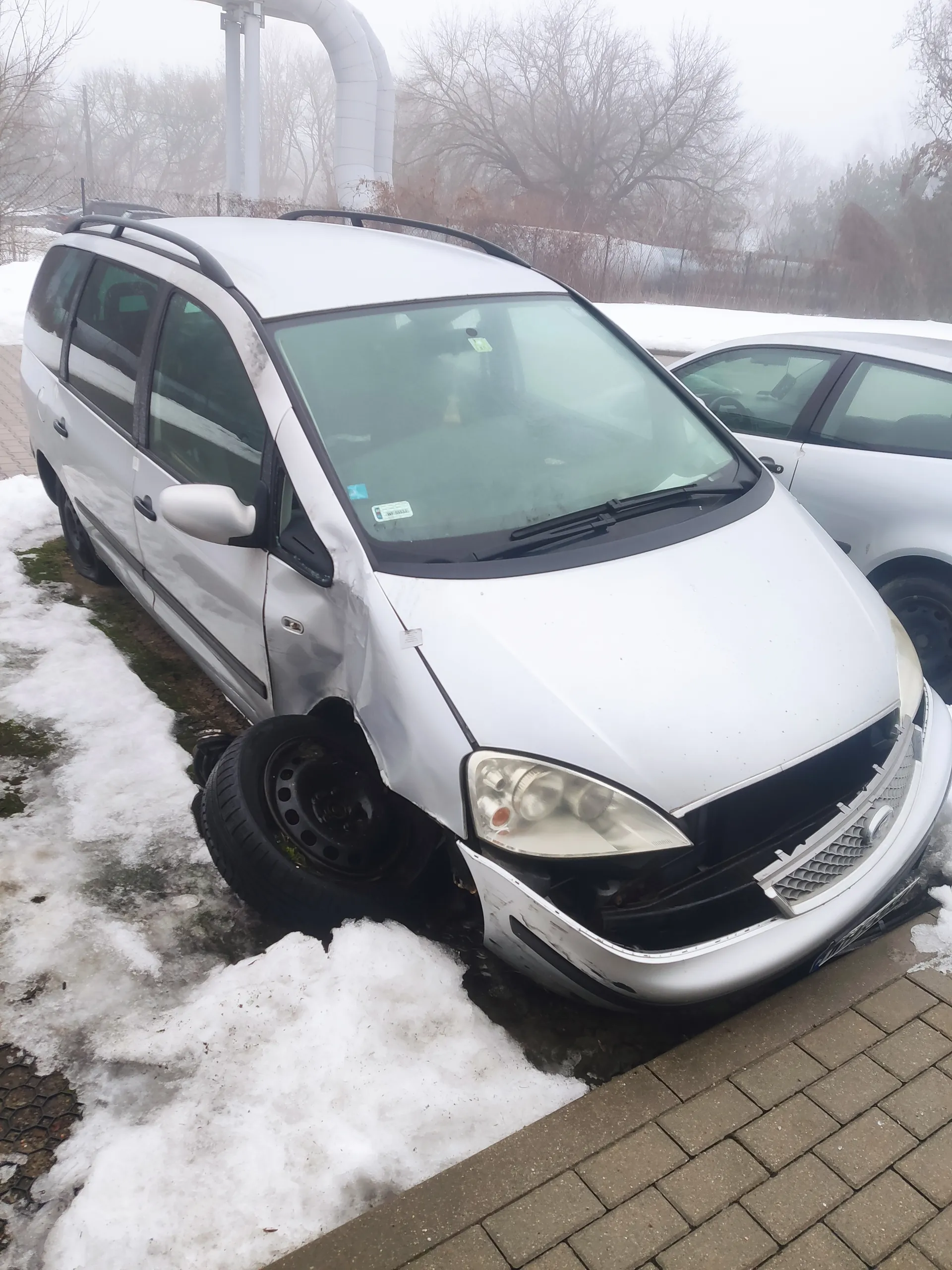 Biały samochód z uszkodzonym przodem zaparkowany na śniegu obok brukowanej nawierzchni. Widoczne uszkodzenia pasa przedniego, w tym zderzaka i reflektora. W tle drugi samochód, drzewa bez liści oraz mglisty krajobraz. Na przedniej szybie znajdują się naklejki.