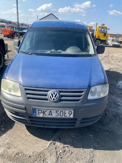 Niebieski samochód osobowy marki Volkswagen, widok z przodu. Tablica rejestracyjna z napisem 'PKA 50L9' oraz oznaczeniem 'PL' po lewej stronie. W tle widoczne są maszyny budowlane, w tym żółty pojazd i pomarańczowy pojazd, na terenie ziemnym pod jasnym niebem z chmurami.