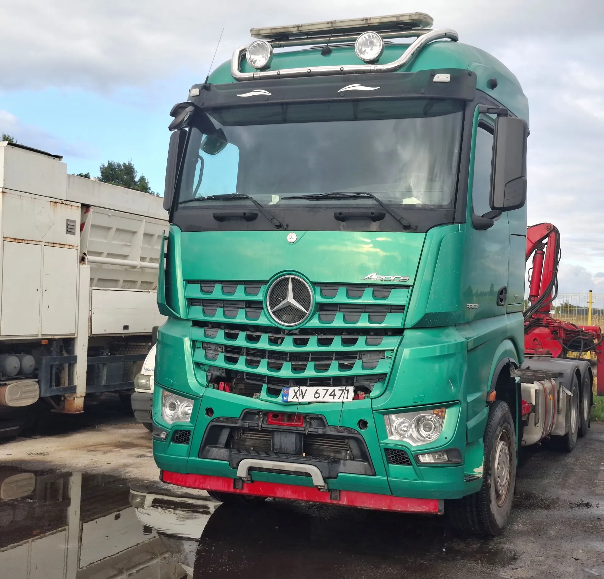 Zielona ciężarówka marki Mercedes-Benz Actros zaparkowana na mokrym placu. Pojazd posiada tablicę rejestracyjną XV 67471. Na dachu kabiny zamontowano dodatkowe reflektory i belkę oświetleniową. Po prawej stronie widoczny jest czerwony żuraw hydrauliczny, a po lewej częściowo widoczna biała naczepa.