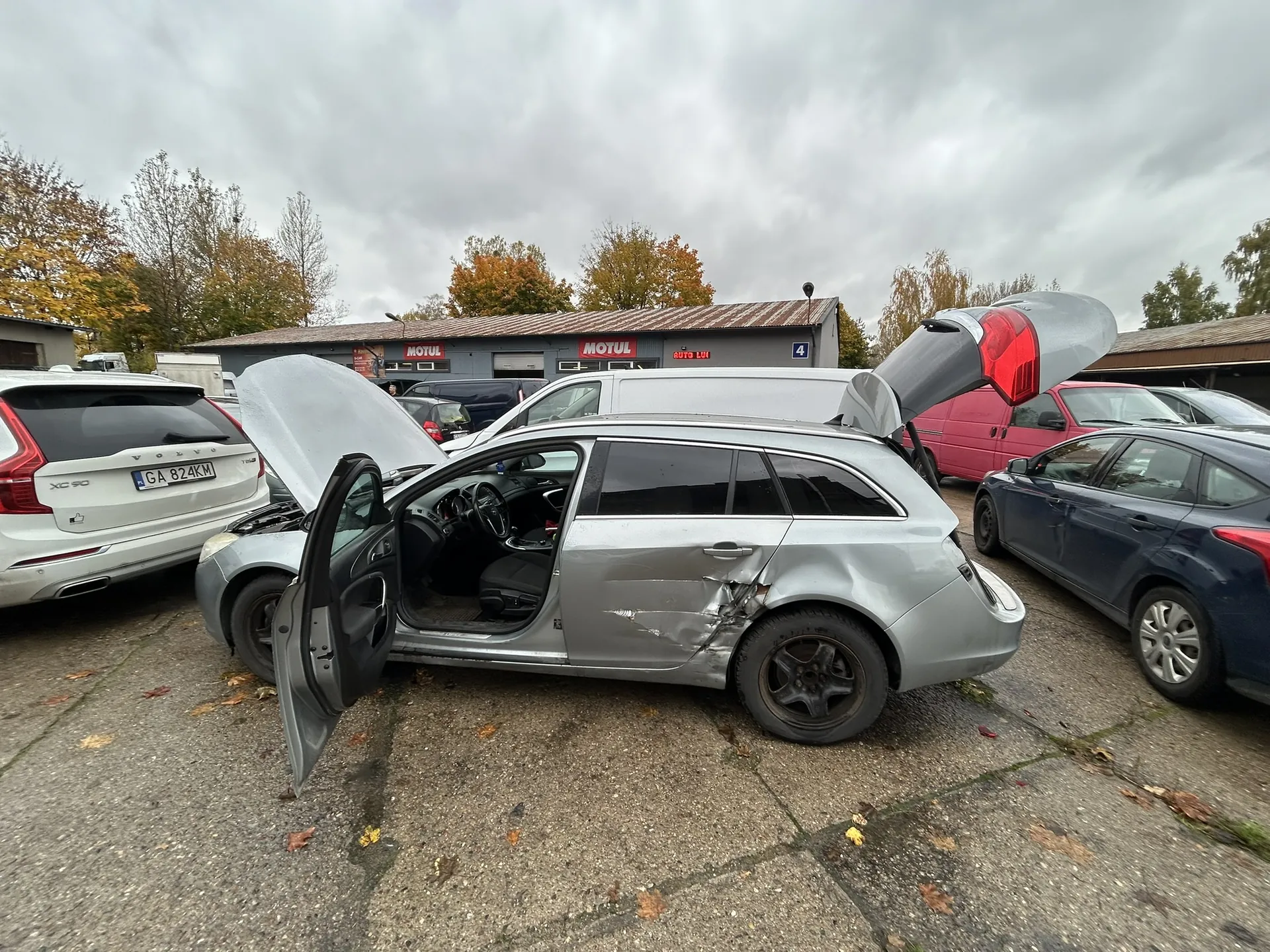 Zdjęcie przedstawia parking z uszkodzonym srebrnym samochodem typu kombi, którego maska, bagażnik i przednia prawa drzwi są otwarte. Po lewej stronie widoczny jest biały samochód Volvo XC90 z tablicą rejestracyjną GA 824KM. W tle znajduje się budynek z czerwonymi napisami MOTUL oraz numerem 4. Wokół stoją inne pojazdy: niebieski samochód osobowy, różowy furgon i inne. Na niebie dominują chmury, a w tle rosną drzewa z jesiennymi liśćmi. Podłoga parkingu pokryta jest suchymi liśćmi.