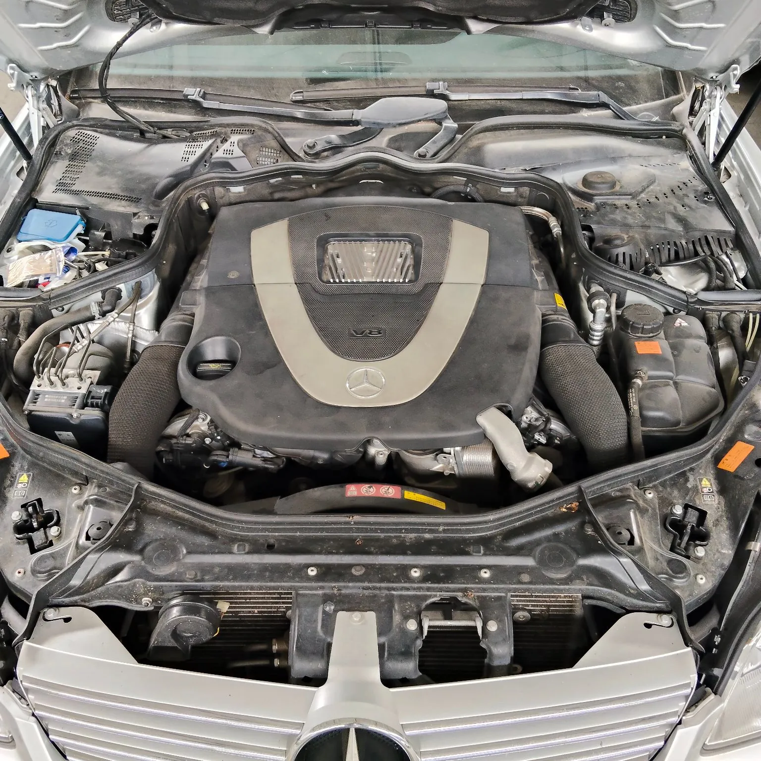 Widok z góry na komorę silnika samochodu marki Mercedes-Benz. Centralnie znajduje się pokrywa silnika w kolorach czarnym i srebrnym z logo gwiazdy Mercedesa na środku oraz oznaczeniem V8. Po bokach widoczne są elementy układu dolotowego, akumulator w niebieskiej obudowie po lewej stronie oraz zbiorniki z płynami eksploatacyjnymi. Na dole zdjęcia widoczny jest fragment srebrnej atrapy chłodnicy z logo marki.