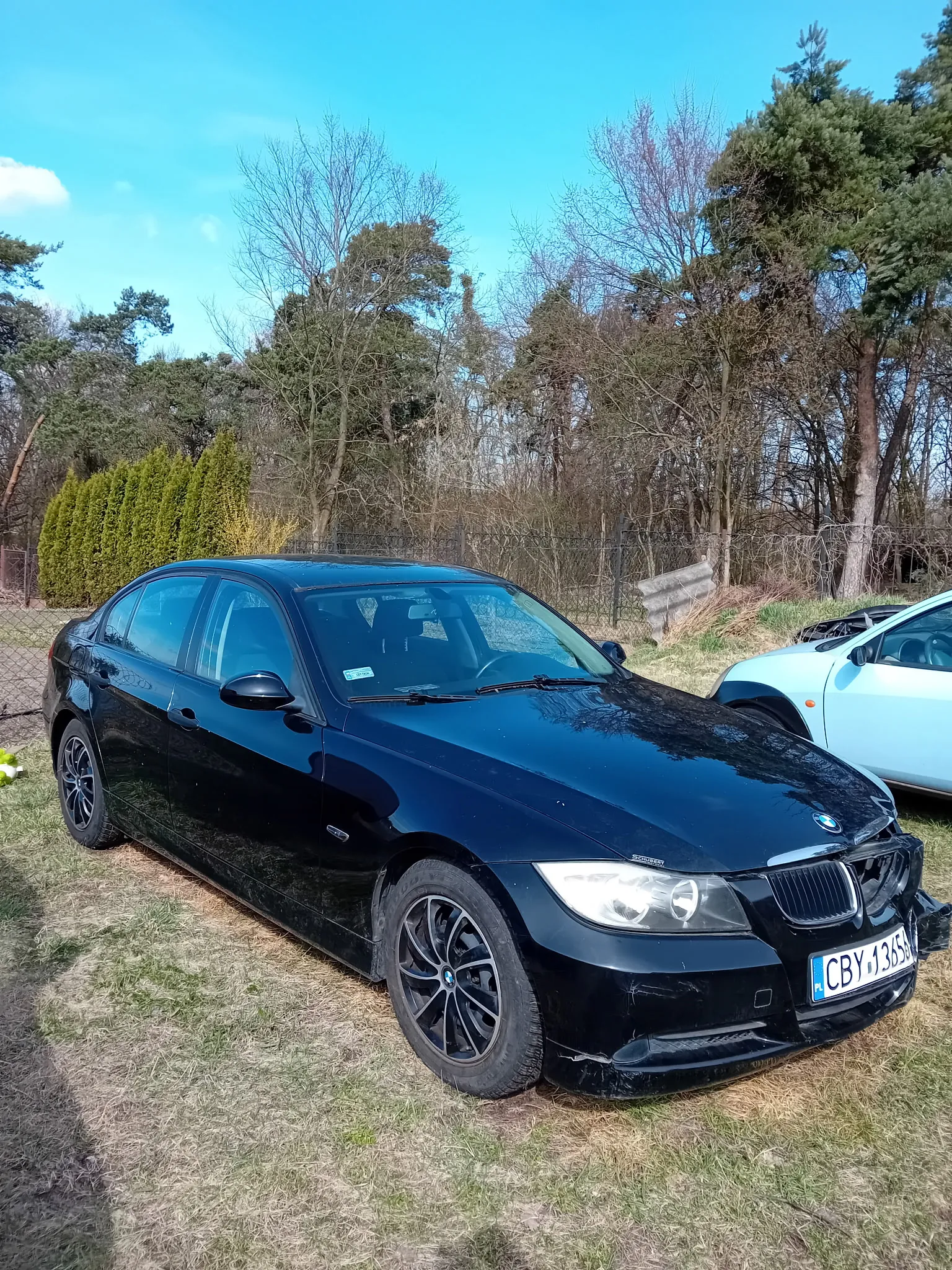 Czarny samochód marki BMW model sedan zaparkowany na trawie. Widoczna tablica rejestracyjna CBY 1365. Obok czarnego BMW znajduje się jasnoniebieski samochód. Tło stanowią drzewa, krzewy, metalowe ogrodzenie oraz błękitne niebo z nielicznymi chmurami. Trawa na obszarze parkowania jest częściowo sucha.