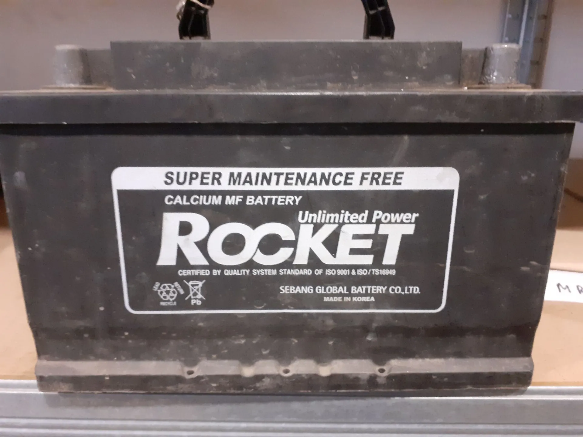 Szary akumulator samochodowy z białą etykietą. Na etykiecie widnieją napisy: 'SUPER MAINTENANCE FREE', 'CALCIUM MF BATTERY', duże logo 'ROCKET' oraz mniejszy napis 'Unlimited Power'. Poniżej znajduje się informacja: 'CERTIFIED BY QUALITY SYSTEM STANDARD OF ISO 9001 & ISO/TS16949', 'SEBANG GLOBAL BATTERY CO., LTD.' oraz 'MADE IN KOREA'. Po lewej stronie widoczne są symbole recyklingu i ołowiu (Pb).