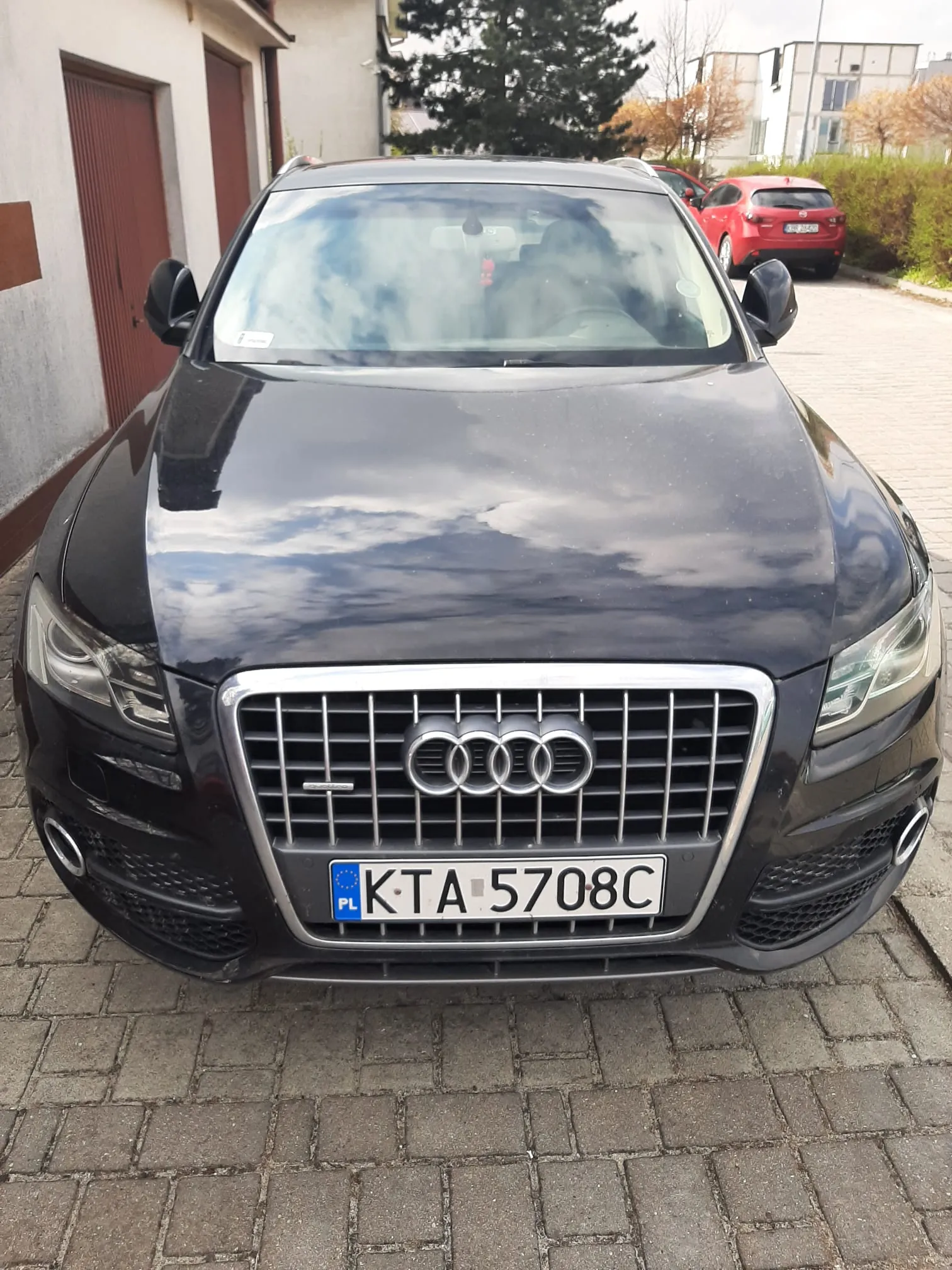 Czarny samochód marki Audi z przodu, z widoczną tablicą rejestracyjną PL KTA 5708C. Samochód stoi na brukowanej powierzchni, w tle budynek z brązowymi drzwiami, drzewa oraz czerwony samochód. Na tablicy rejestracyjnej widoczny jest niebieski pas z literą PL i symbolu Unii Europejskiej. Chmury odbijają się w szybie i kapotie pojazdu.