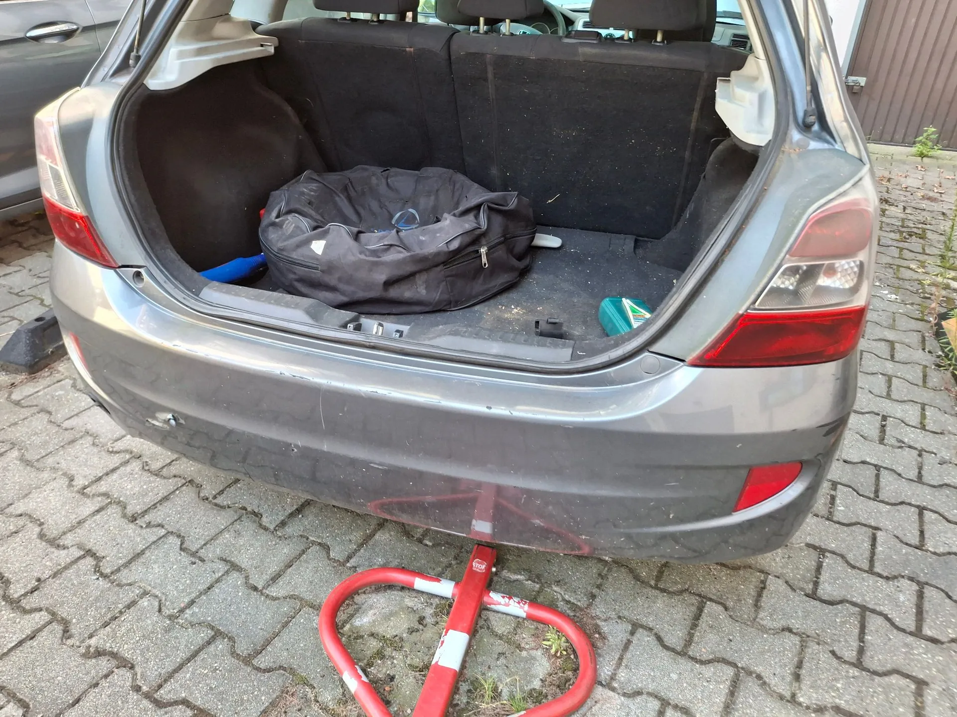 Otwarty bagażnik szarego samochodu zaparkowanego na kostce brukowej. Wewnątrz bagażnika znajduje się duża, czarna torba sportowa, niebieski przedmiot oraz mała zielona butelka. Pod tylnym zderzakiem samochodu widoczna jest czerwona, metalowa konstrukcja przypominająca stojak lub element zabezpieczający.