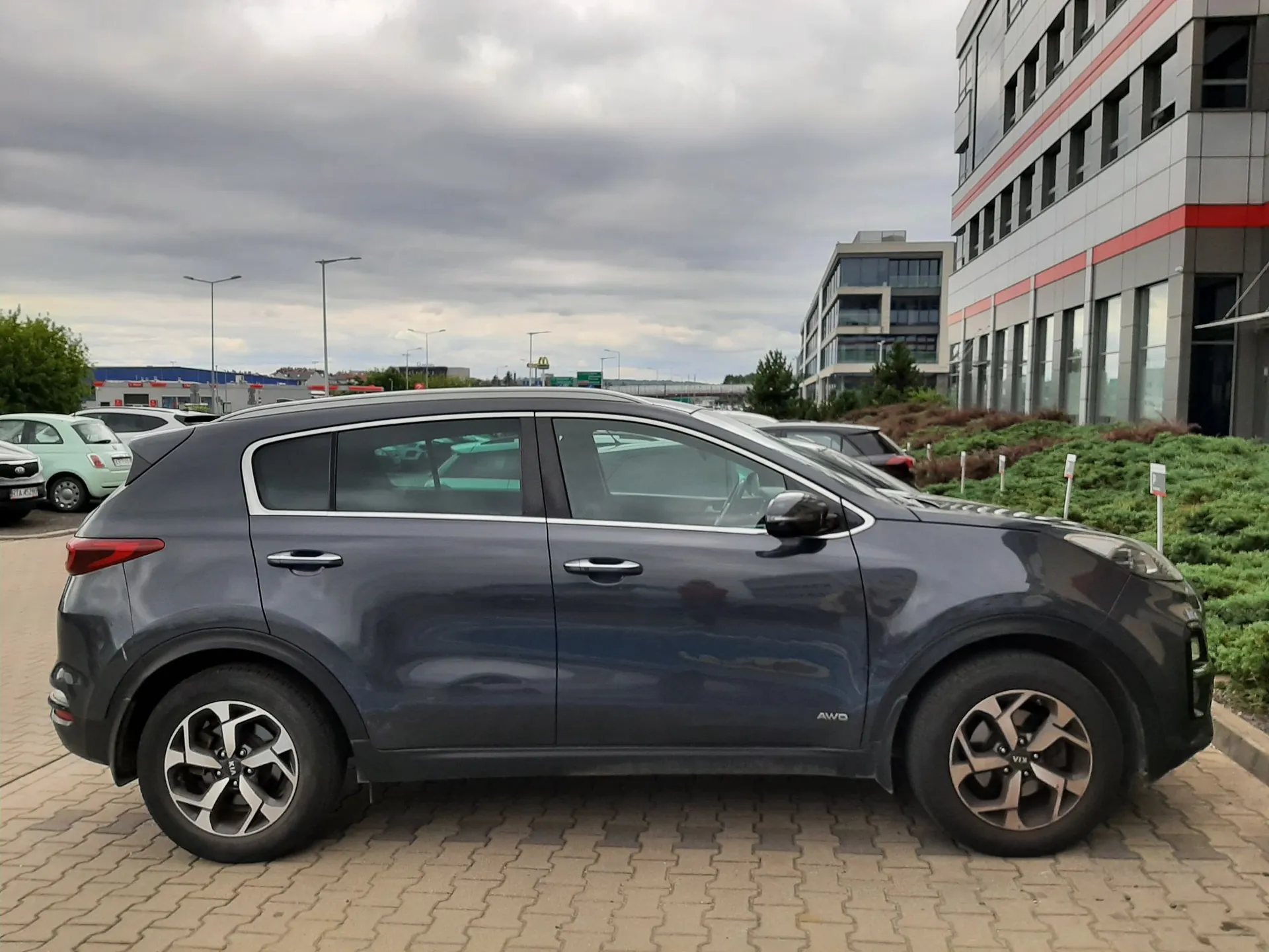 Szary samochód typu SUV zaparkowany bokiem na kostce brukowej. W tle widoczne są inne zaparkowane pojazdy, nowoczesny biały budynek biurowy z czerwonymi akcentami oraz zachmurzone niebo.