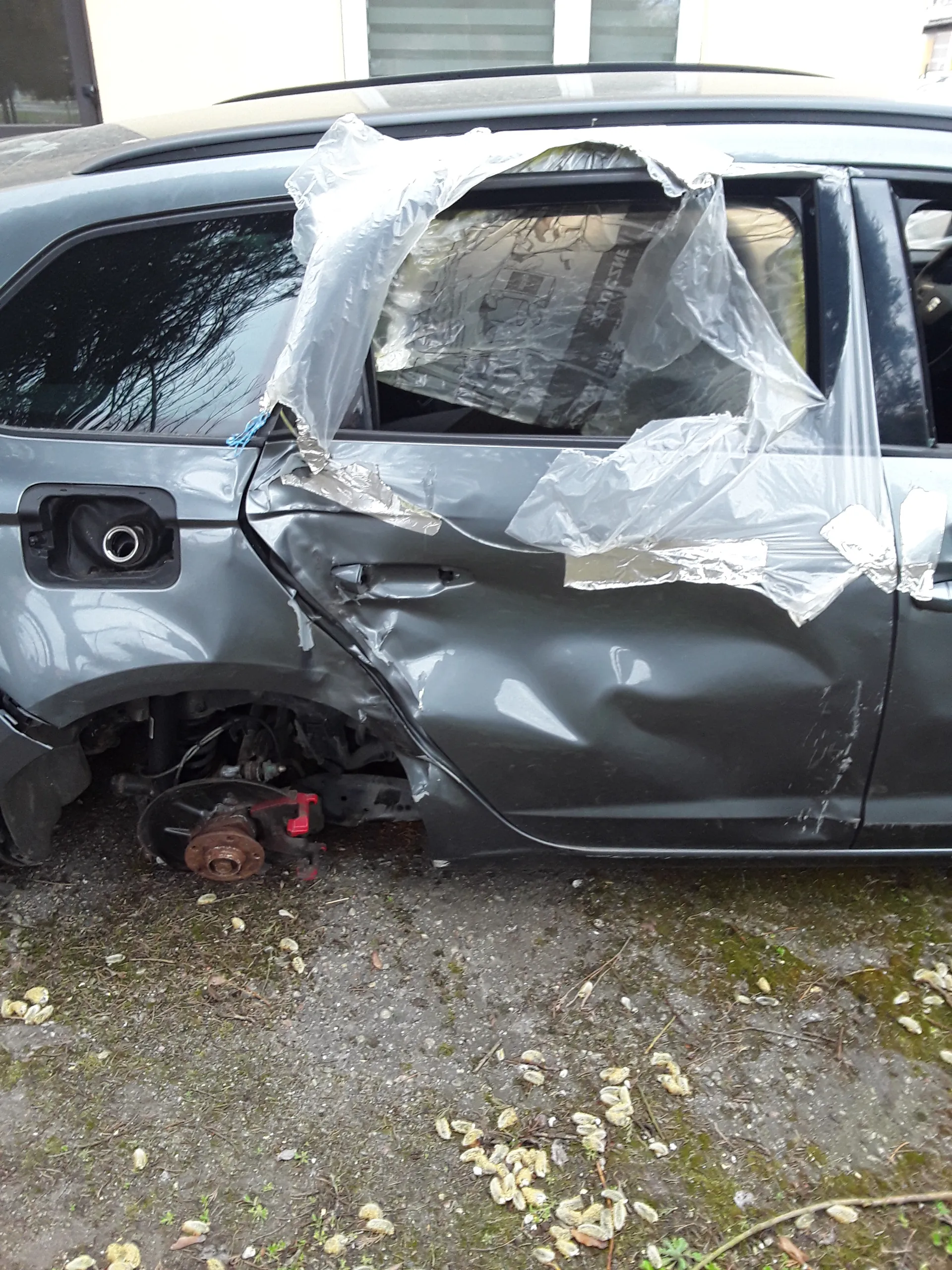 Zniszczony szary samochód z uszkodzonym przodem, zdeformowaną maską i brakującym przednim kołem. Widoczny zacisk hamulcowy oraz tarcza hamulcowa. Przednią szybę częściowo zakrywa przezroczysta folia z widocznym napisem 'SUN'. Samochód zaparkowany na podłożu z żwirem i mchem, w tle fragment budynku z oknami.