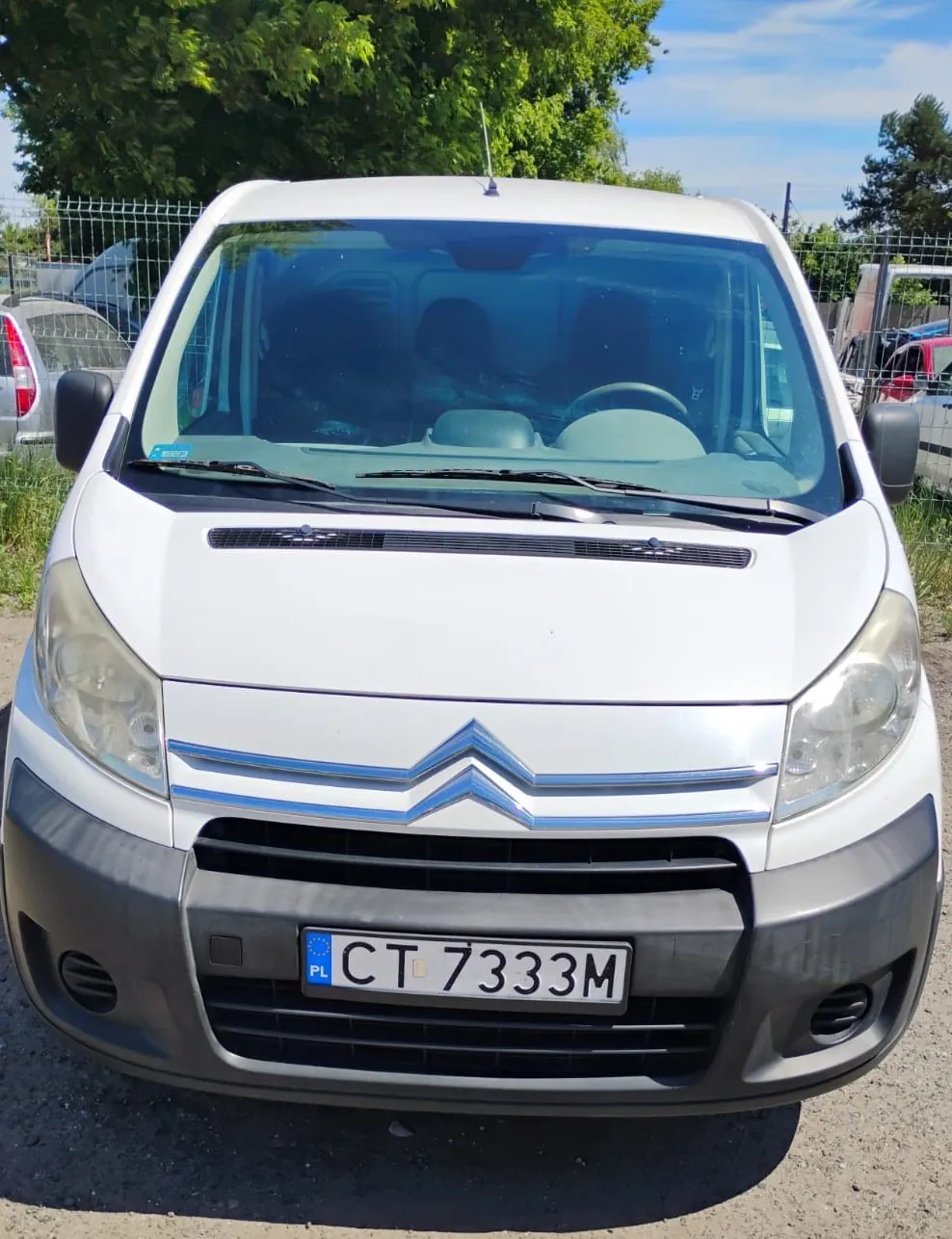 Widok z przodu białego samochodu dostawczego marki Citroën. Pojazd posiada czarny zderzak i polską tablicę rejestracyjną o numerze CT 7333M. W tle widoczne są zielone drzewa, niebieskie niebo oraz ogrodzenie z siatki.