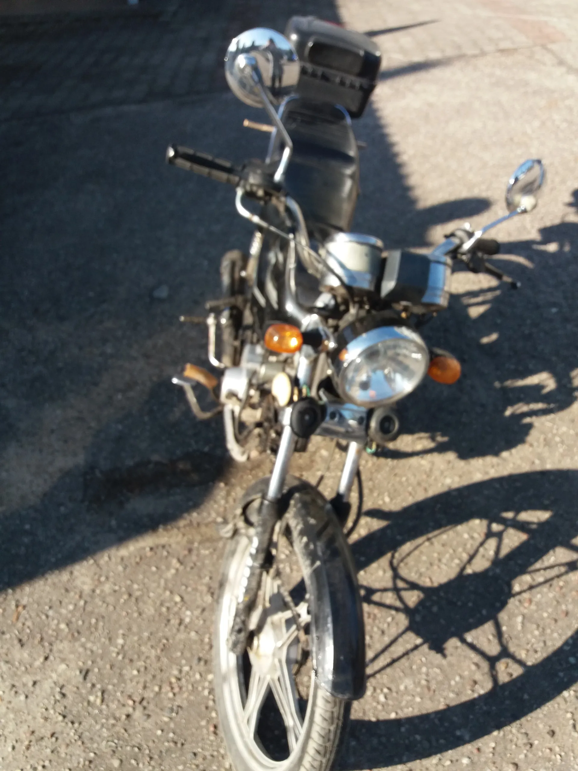 Motocykl na asfaltowej powierzchni, widziany z góry. Czarna siedzisko, chromowane elementy, reflektor, pomarańczowe kierunkowskazy, lusterka boczne i przednie koło. W tle widoczne cienie motocykla oraz osób.