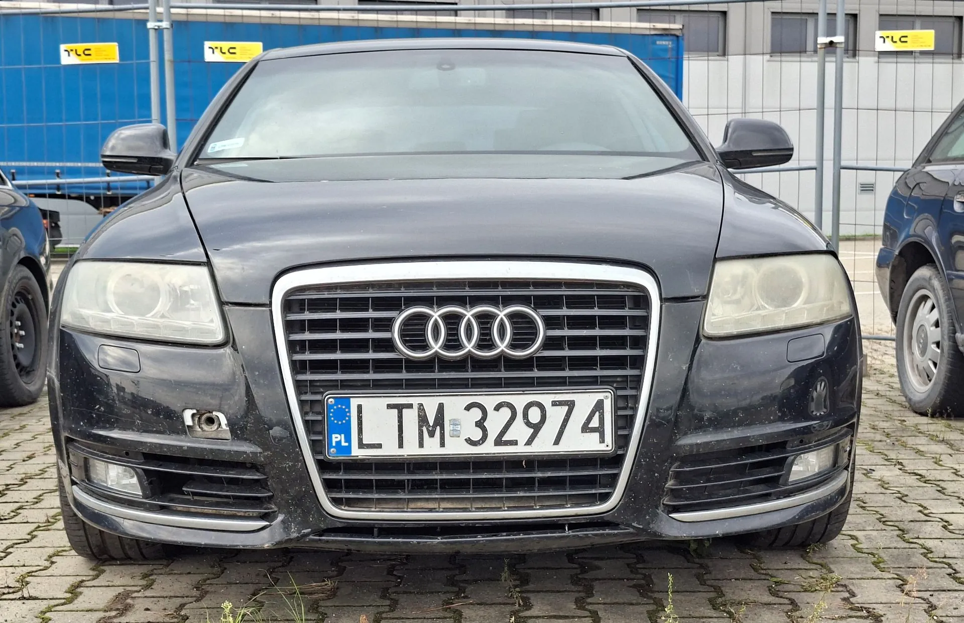 Zdjęcie przodu czarnego samochodu marki Audi zaparkowanego na kostce brukowej. Samochód ma polską tablicę rejestracyjną o numerze LTM 32974. W tle widoczny jest niebieski budynek z żółtym logo TLC oraz fragment innego ciemnego pojazdu po prawej stronie.