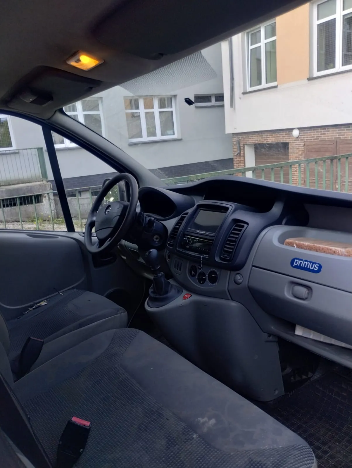Wnętrze samochodu z widoczną deską rozdzielczą, kierownicą, siedzeniami i ekranem centralnym. Na desce rozdzielczej znajduje się niebieski napis 'primus'. W przednim oknie widoczne są budynki z oknami i zielonym ogrodzeniem. Na desce rozdzielczej leży kromka chleba.