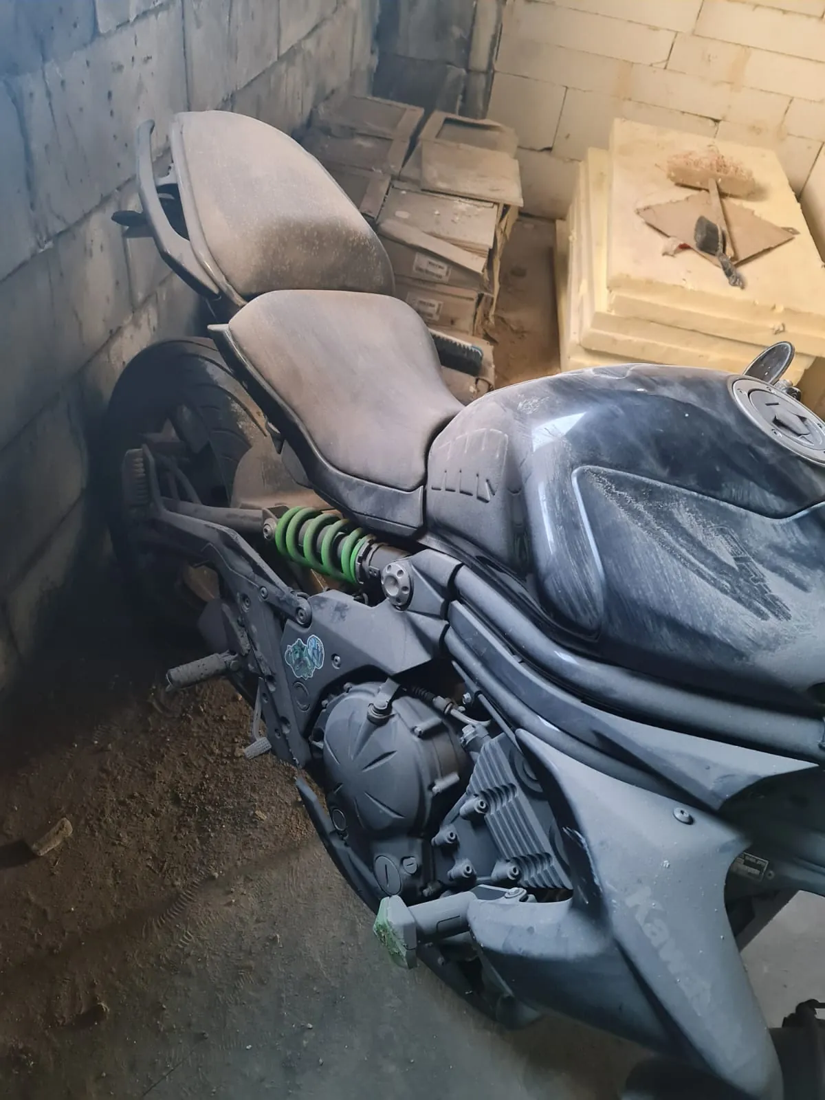 Czarny motocykl Kawasaki w stanie zapylonym, zaparkowany w pomieszczeniu z betonowymi blokami. Widoczne są zielone sprężyny zawieszenia, siedzenie oraz zbiornik paliwa pokryte kurzem. W tle stosy kartonów i desek, na których leżą różne przedmioty. Podłoga z betonu z widocznymi śladami brudu.