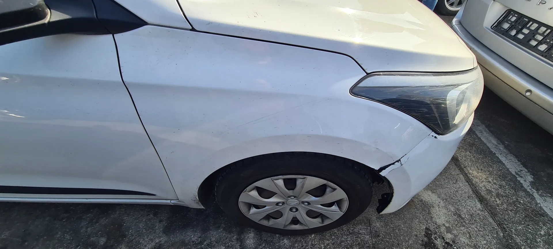 Biały samochód widziany z przodu po prawej stronie, z widocznym uszkodzeniem nad przodem pasa przedniego koła. Widoczne są reflektor, felga aluminiowa oraz fragment srebrnego samochodu po prawej stronie. Tło stanowi asfaltowa powierzchnia parkingu z białymi liniami.