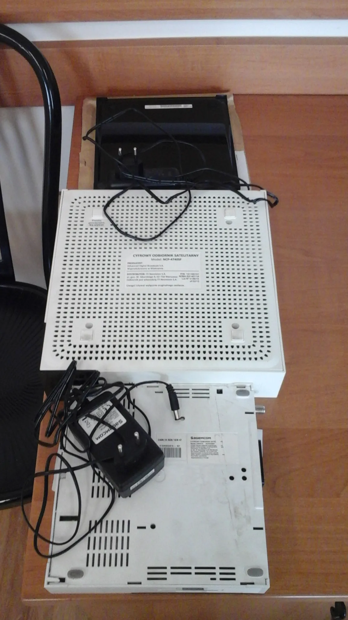 Biały cyfrowy odbiornik satelitarny model KX-4740SF z siatką wentylacyjną i etykietą z tekstem. Obok znajduje się drugi biały urządzenie z czarnymi kabelkami i czarny zasilacz z napisem Sagemcom. Wszystko leży na drewnianym blacie. Widoczne są również czarne przewody i czarna obudowa urządzenia w tle.