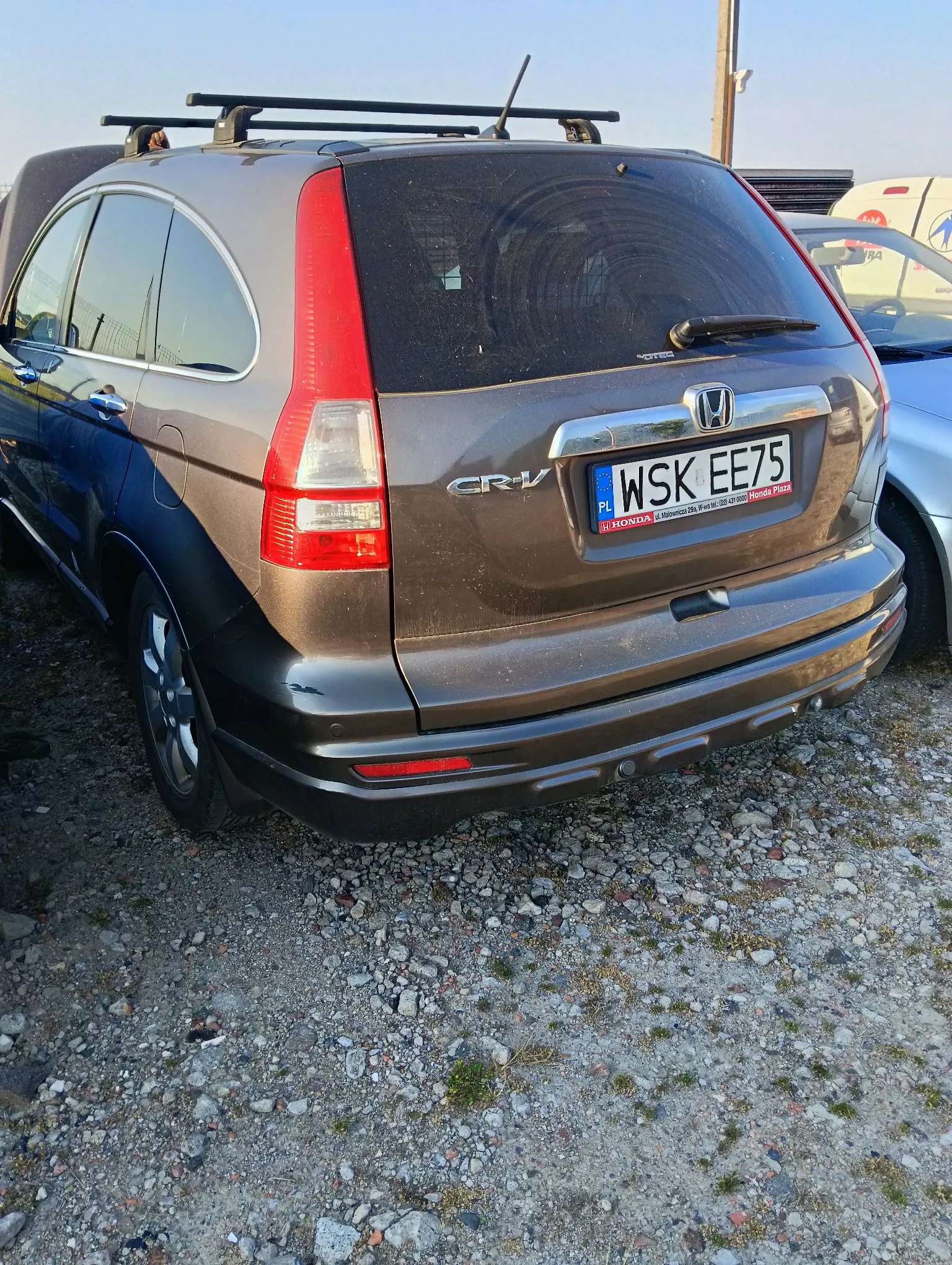 Tylny widok brązowego samochodu marki Honda CR-V zaparkowanego na żwirowym podłożu. Pojazd posiada zamontowany czarny bagażnik dachowy. Na klapie bagażnika widoczna jest tablica rejestracyjna o numerze WSK EE75 oraz logo Hondy i oznaczenie modelu CR-V. Obok stoi fragment innego, niebieskiego samochodu.